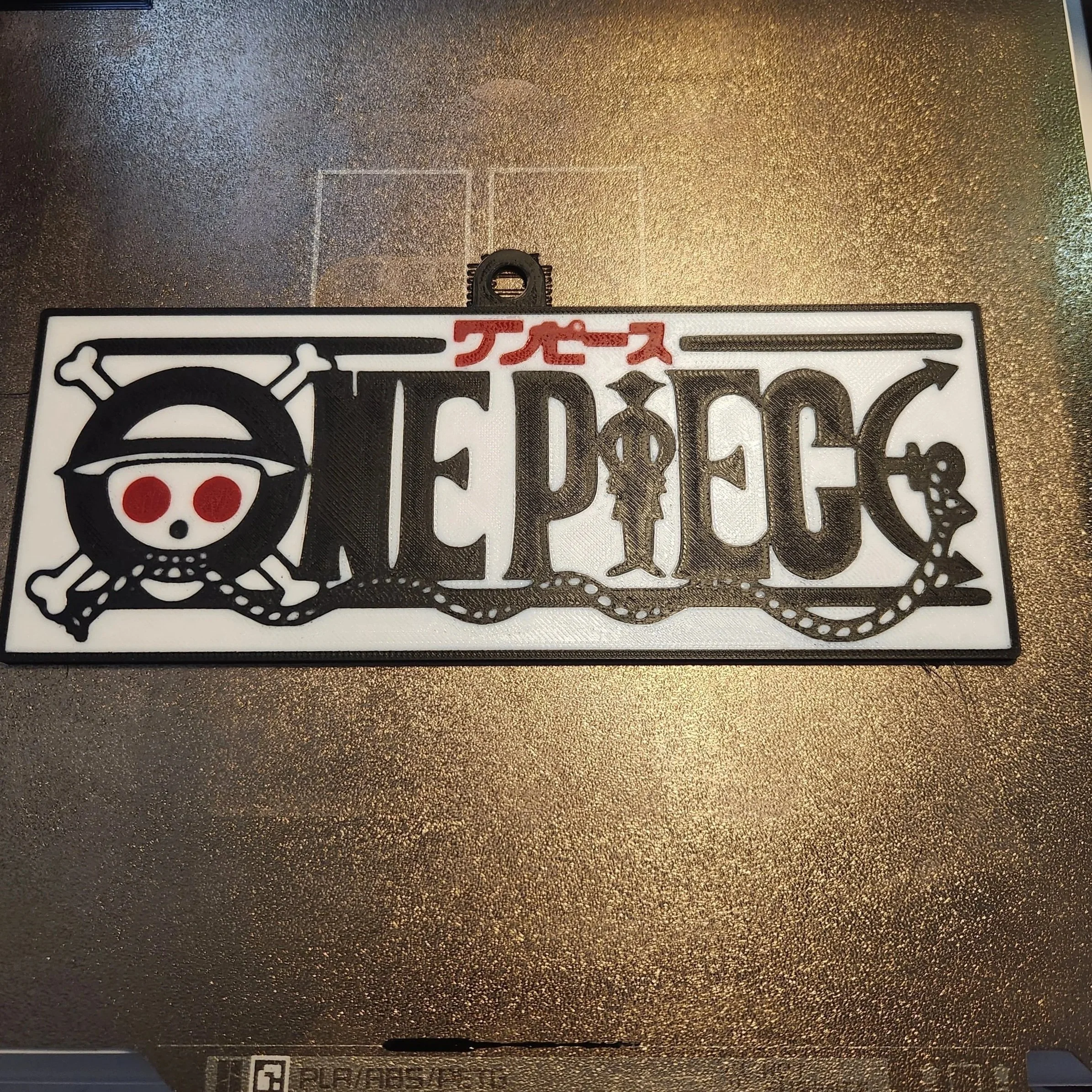 Close-up of a metallic surface with a black carabiner clip attached. Part of a sticker featuring a stylized animal face, with Japanese characters in red above it. The background is textured with a dark color.