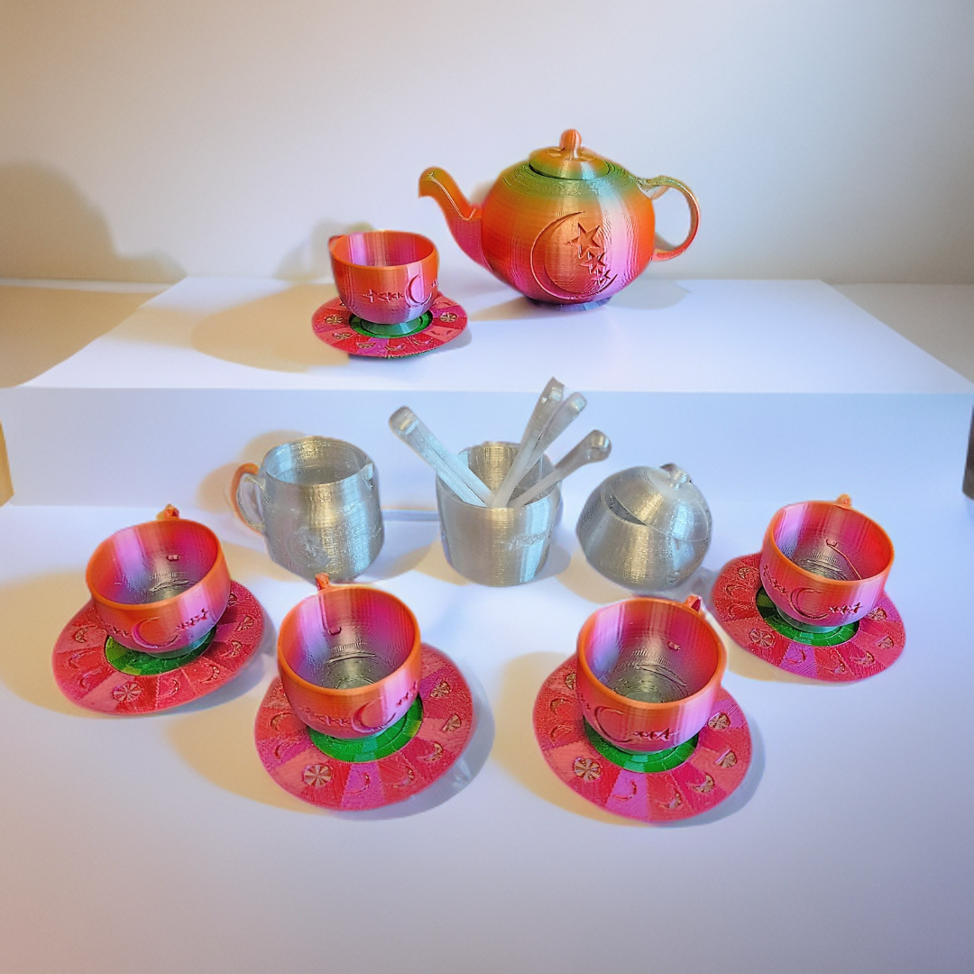 Colorful tea set with a teapot, teacups, saucers, and utensils, made from metallic material, displayed on white surfaces.