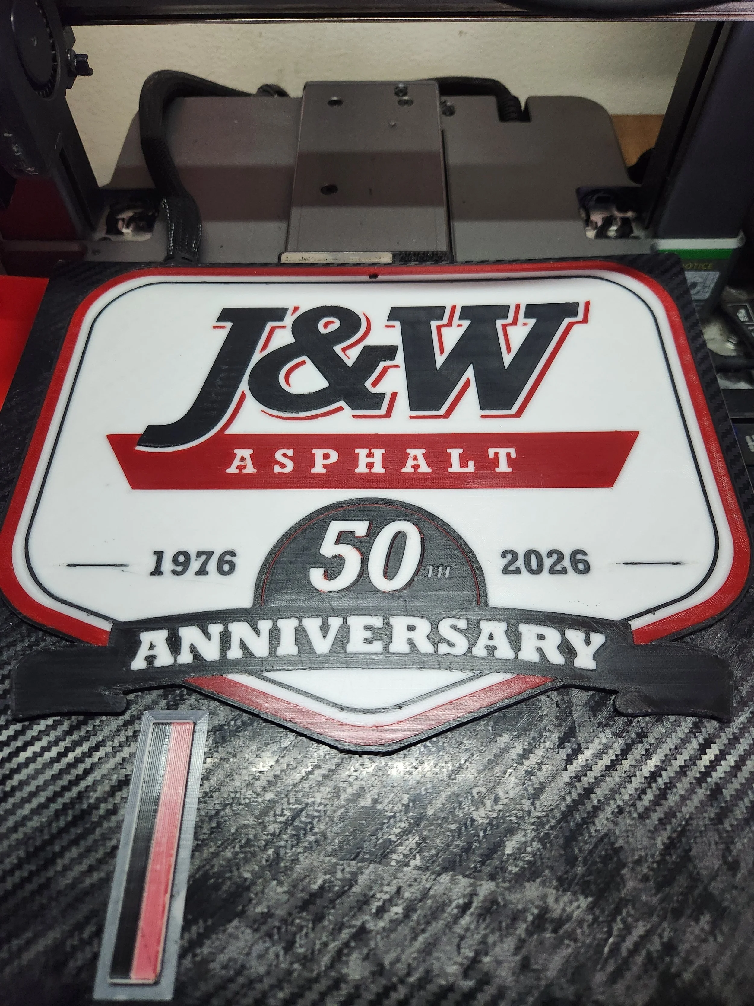 A racing-style license plate celebrating the 50th anniversary of J&W Asphalt from 1976 to 2026, resting on a textured surface.