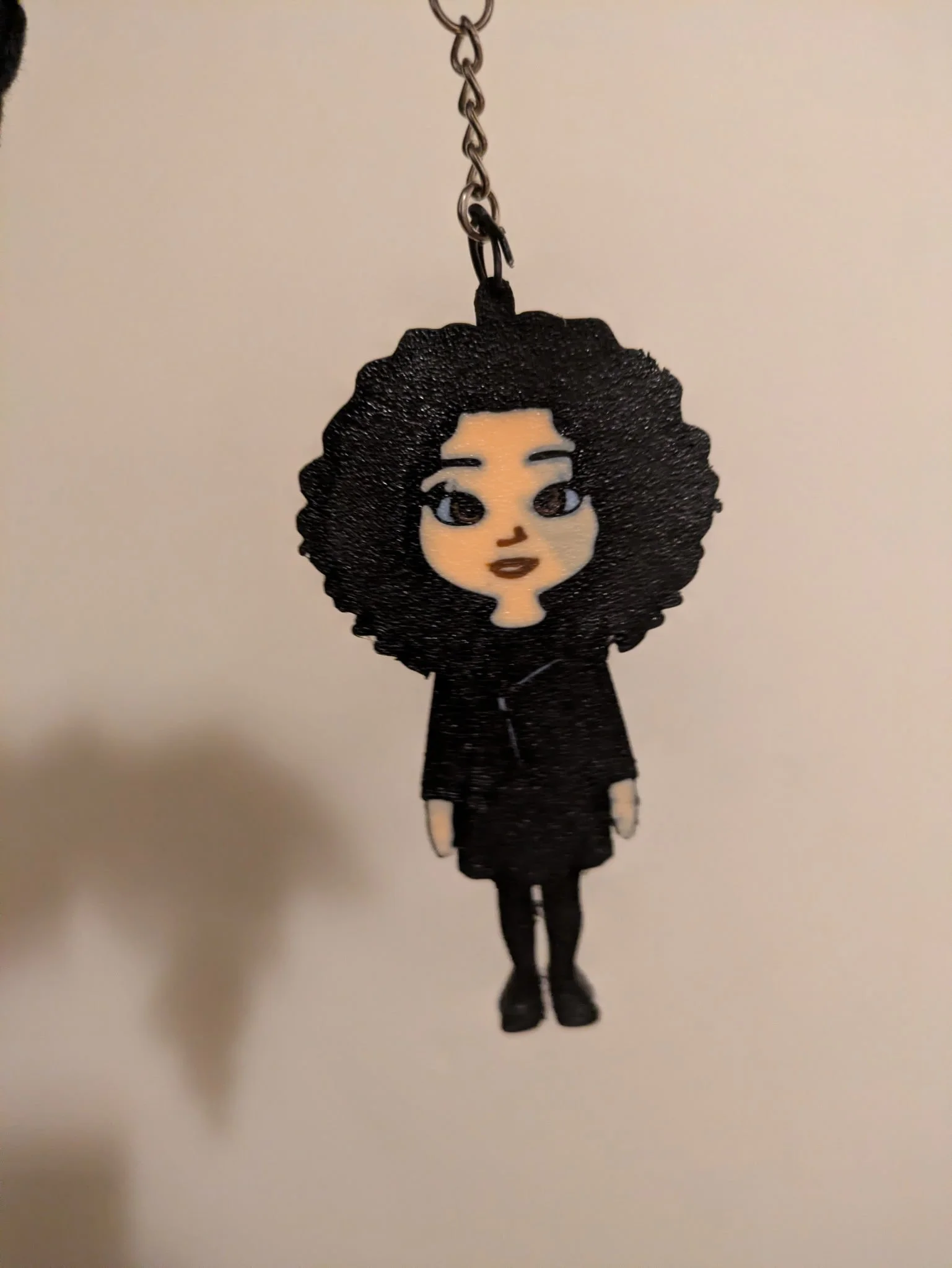 A small figurine of a woman with curly black hair, wearing a black dress and black shoes, hanging from a chain.