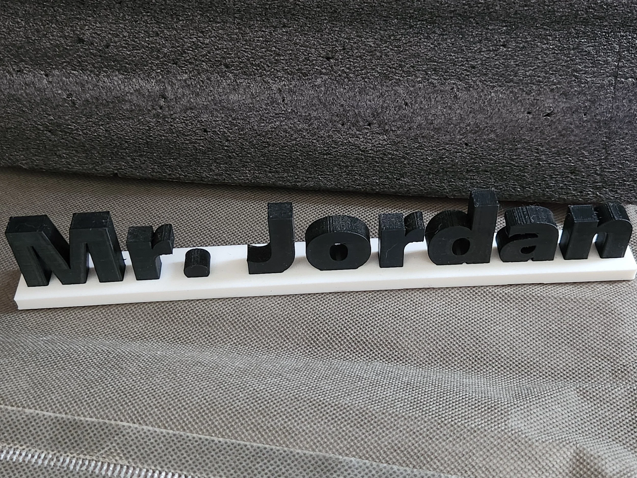 Close-up of black plastic letters spelling 'Morale' on a white base, placed on a textured gray surface with a dark gray fabric backdrop.