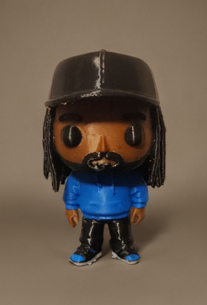 A vinyl figure of a man with dreadlocks, wearing a black cap, blue hoodie, black pants with white details, and sneakers, standing against a plain background.