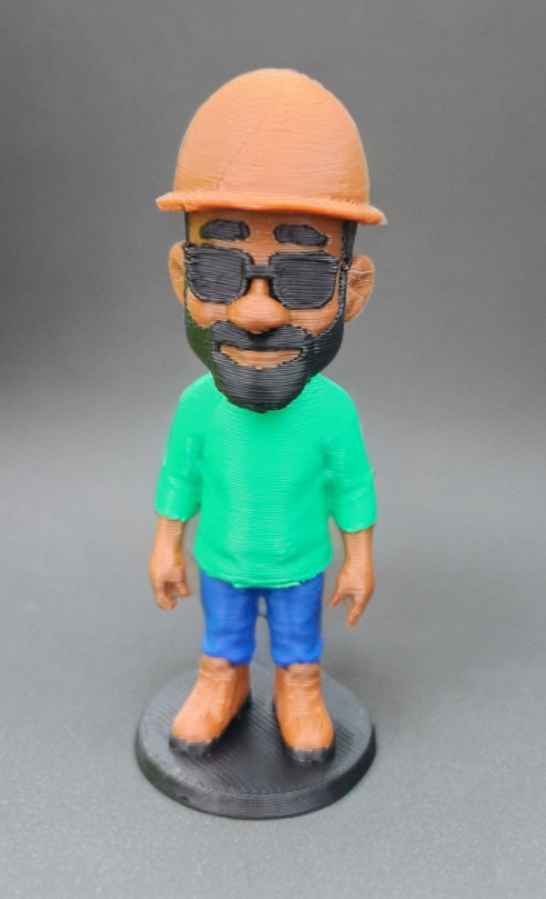 Colorful 3D-printed figurine of a man with a beard, wearing sunglasses, an orange cap, a green shirt, blue pants, and brown shoes, standing on a black circular base.