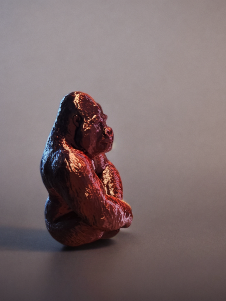 A small, sculpted figure of a gorilla sitting on a plain dark surface with a plain light grey background.