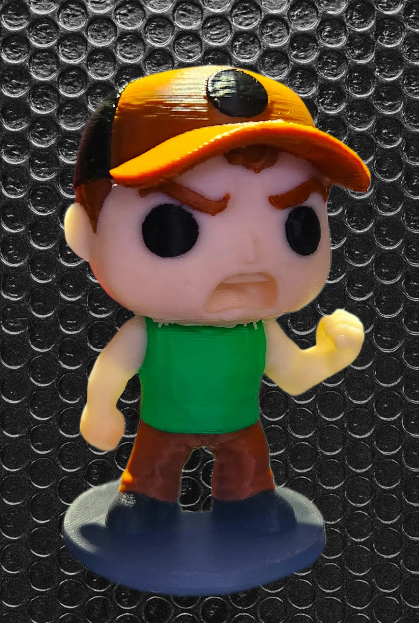 A figurine of a cartoon character with a yellow, black, and orange hat resembling a ladybug, a pink face with large black eyes, wearing a green shirt, brown pants, and dark shoes, standing on a circular gray base against a black bubble wrap backgroun