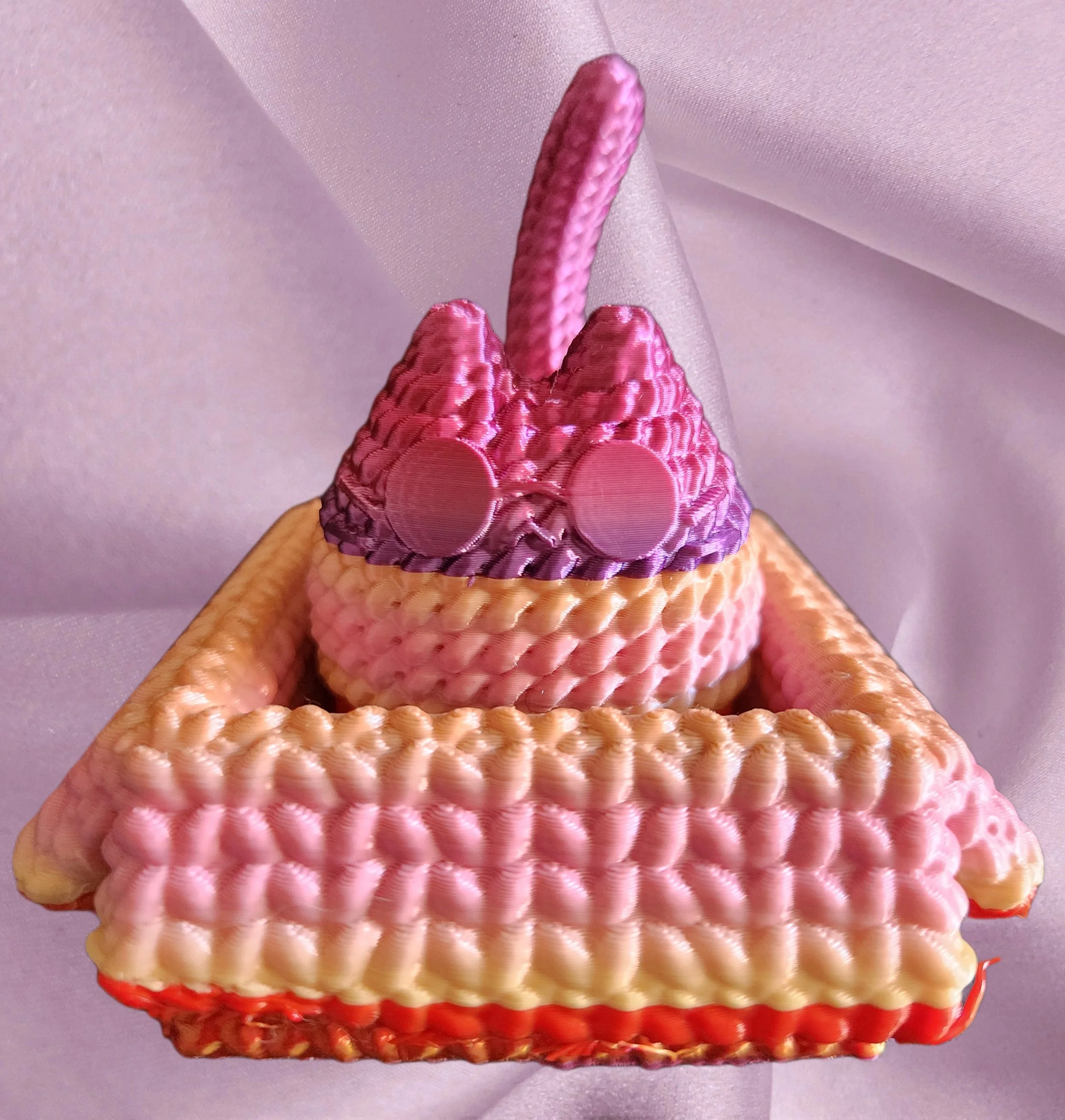A close-up view of a pink and red layered toy or cake with a textured, bubble-like surface, placed on a purple fabric background.