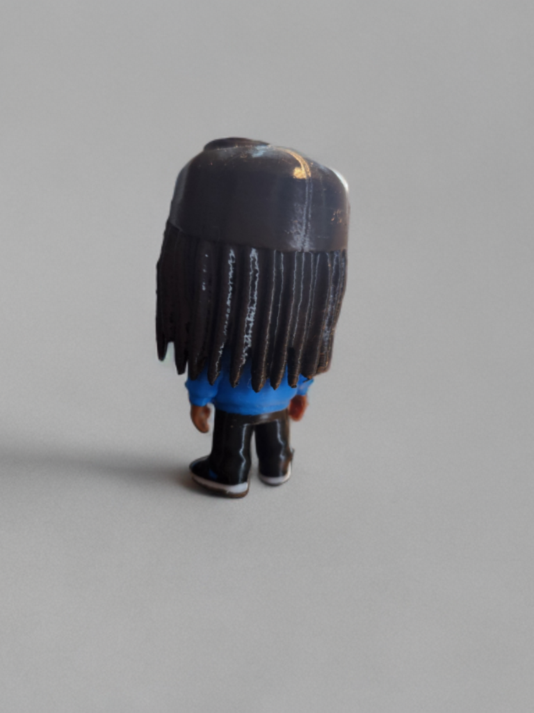 Back of a small figurine with long black hair, wearing a blue shirt, black pants, and sneakers, standing on a plain grey surface.