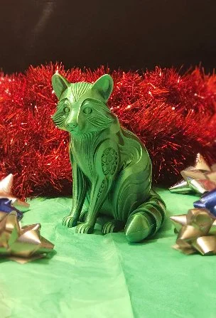 A green, zebra-striped cat figurine sitting on a green surface, with red tinsel and gold and silver bow decorations surrounding it.
