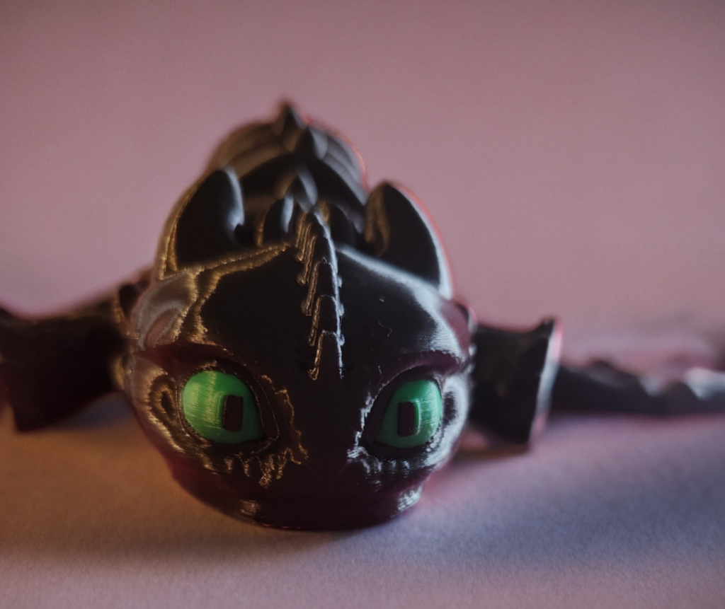 Close-up of a black dragon plush toy with green eyes on a pink surface against a pink background.