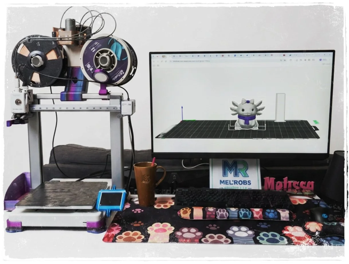 3D printer with filament spools, a computer monitor displaying a 3D model of a cute, small creature with multiple limbs and a scarf, a chessboard, a mug, a blue manual controller, and colorful paw print-themed desk mat.