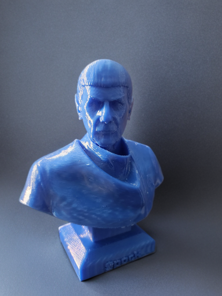 Blue 3D-printed bust of a man with a stern expression, short hair, and wearing a shirt, mounted on a rectangular stand.
