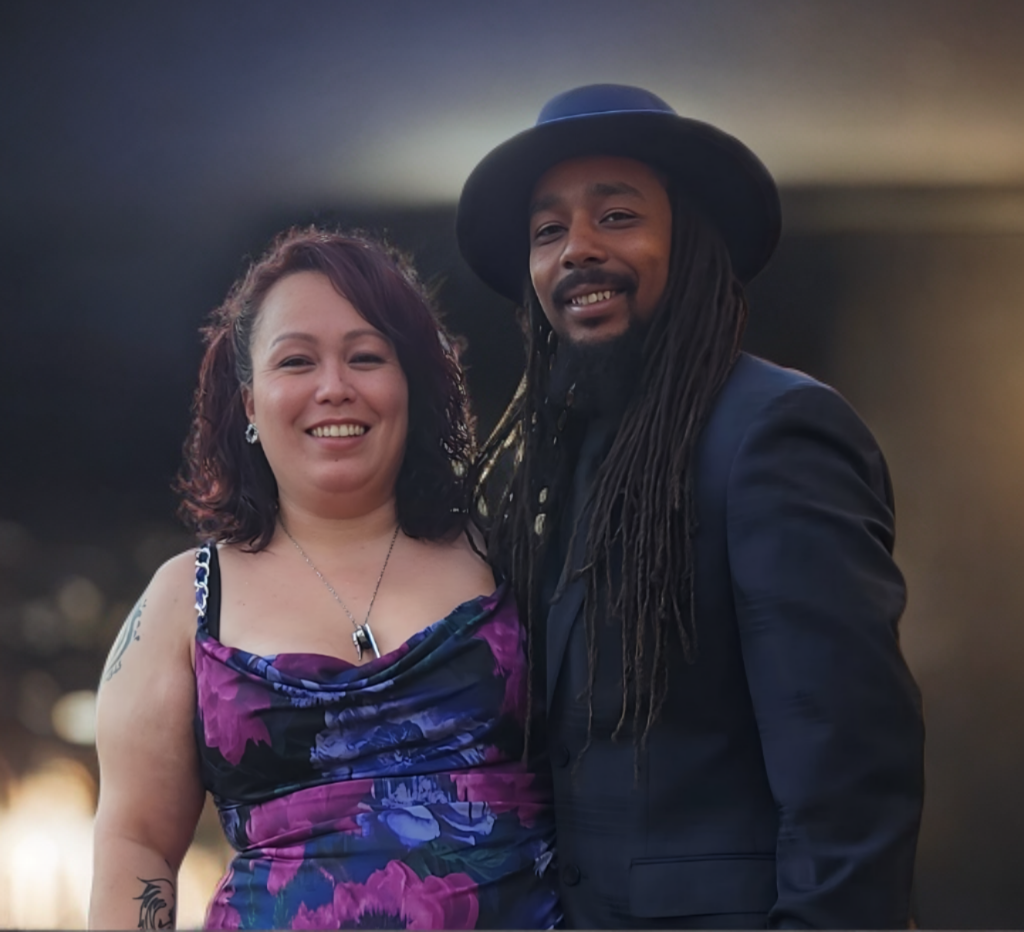A smiling woman with tattoos on her upper arm and sleeveless dress with purple, pink, and blue floral pattern stands next to a smiling man with long dreadlocks, wearing a black hat and dark suit, in a dimly lit setting.