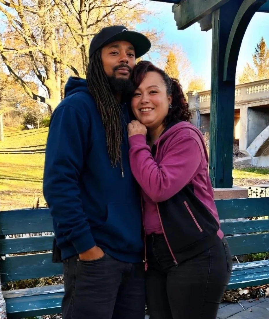 A man and woman standing close together on a park bench, smiling, in an outdoor setting with trees and a bridge in the background.