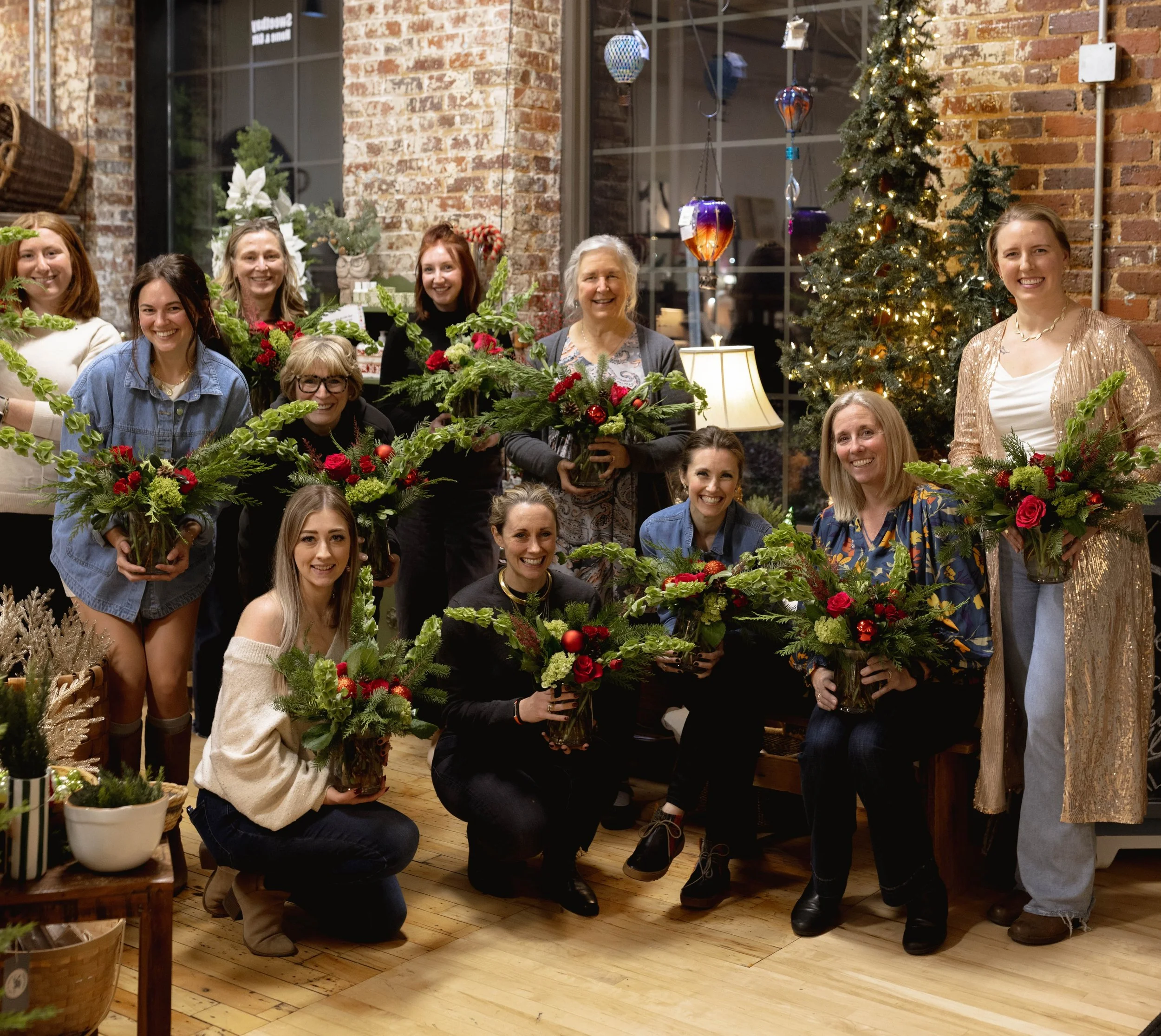 Group of women in a decorated indoor space holding holiday floral arrangements, with Christmas trees and ornaments in the background.