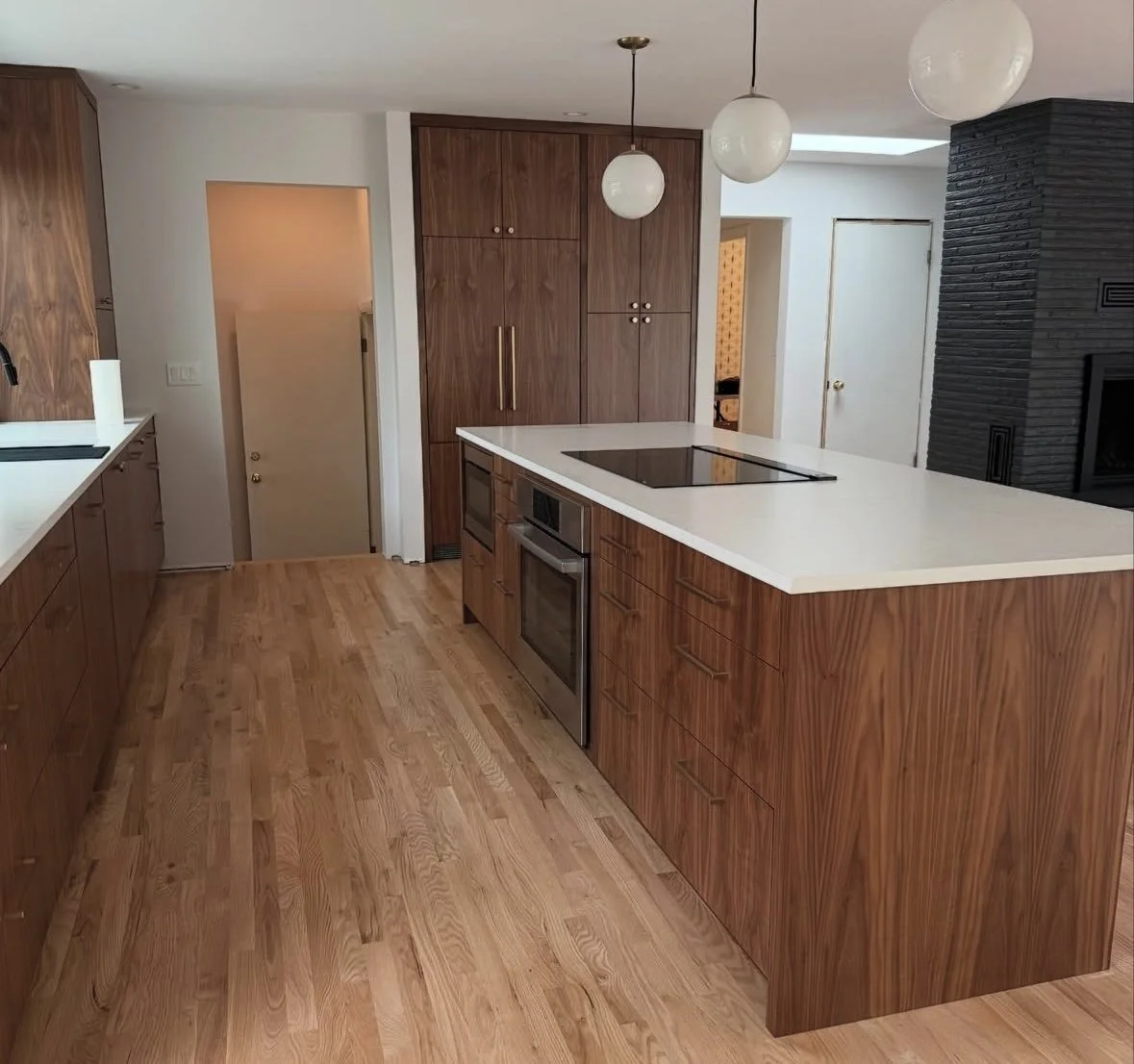 Modern kitchen with wooden cabinets, white countertops, an electric cooktop on the island, pendant lighting, and a black brick fireplace in the background.