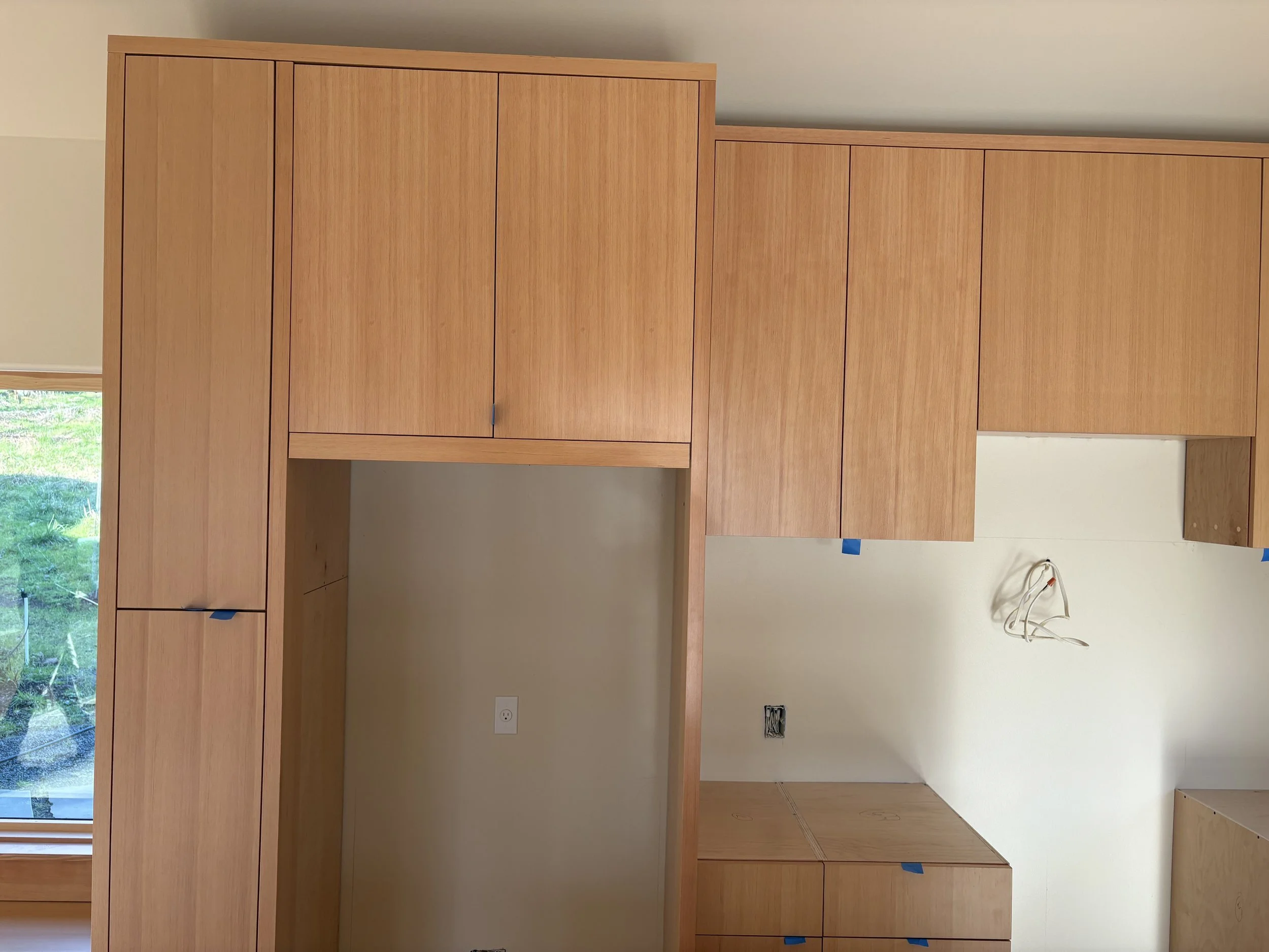 Interior view of a room under construction with light wood cabinets and drawers, a window showing greenery outside, and exposed electrical wires on the wall.