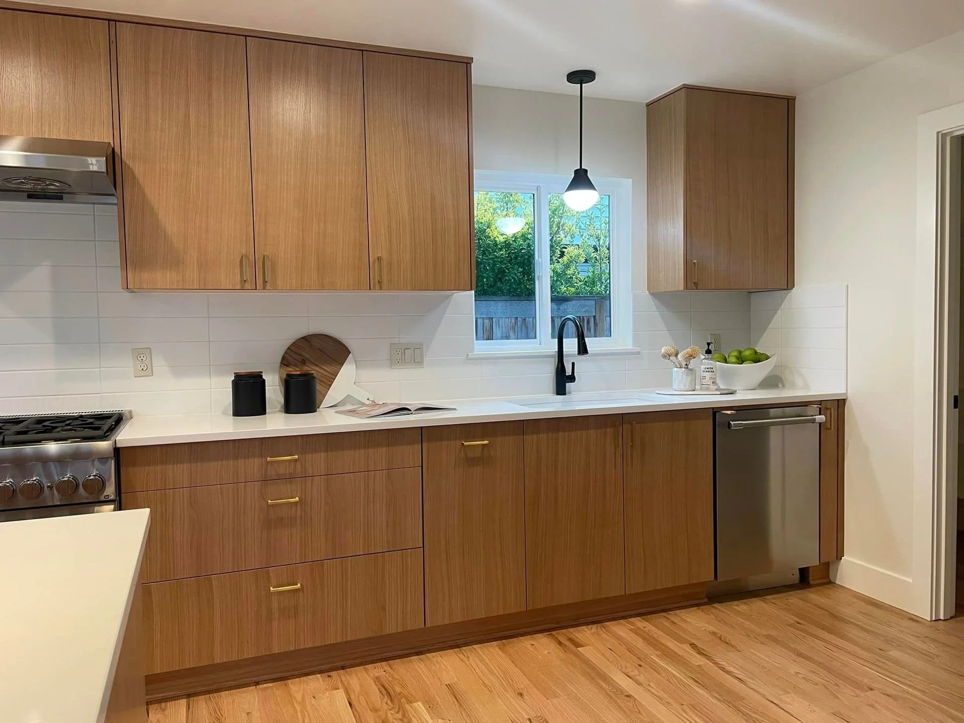Kitchen with wood cabinets, white countertop, black faucet, window with greenery outside, stainless steel dishwasher, and stove, with decorative items including bowls and bottles.