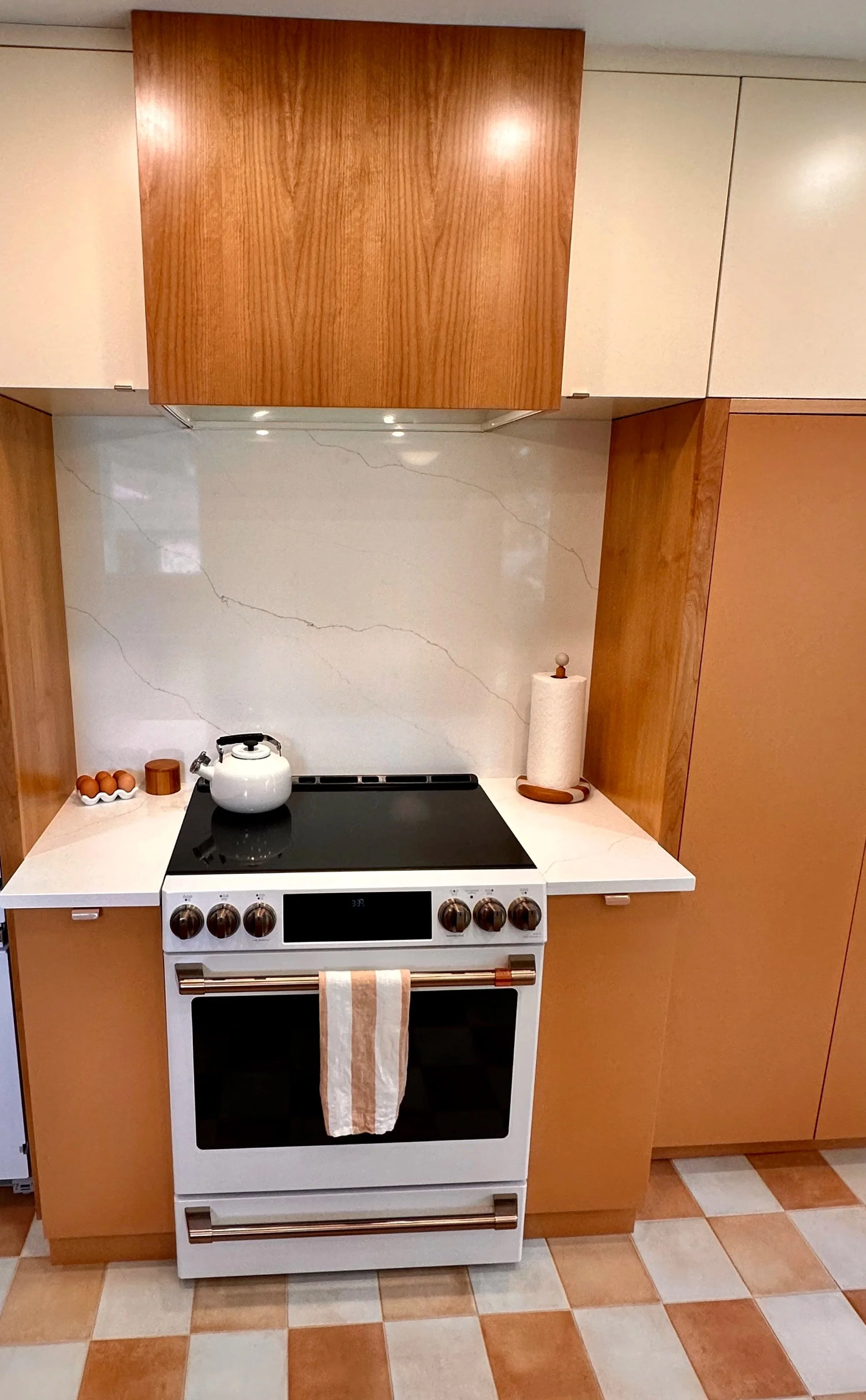 Kitchen stove with a teapot on top, surrounded by wooden cabinets, a roll of paper towels, and eggs on the counter.