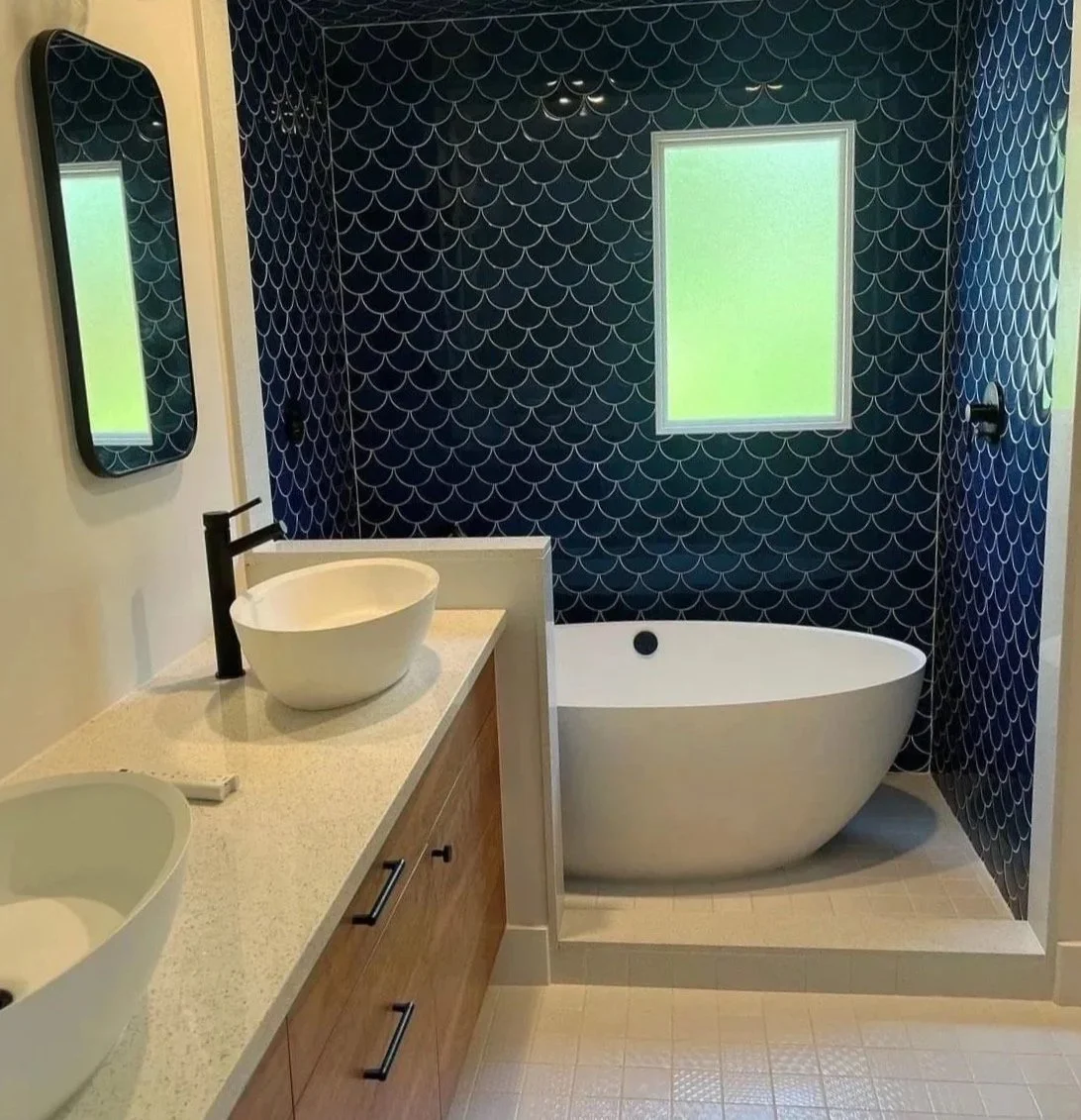Modern bathroom with a white free-standing bathtub, a dual sink vanity with vessel sinks, a large mirror, and navy blue tiled walls with a fish scale pattern and a window.
