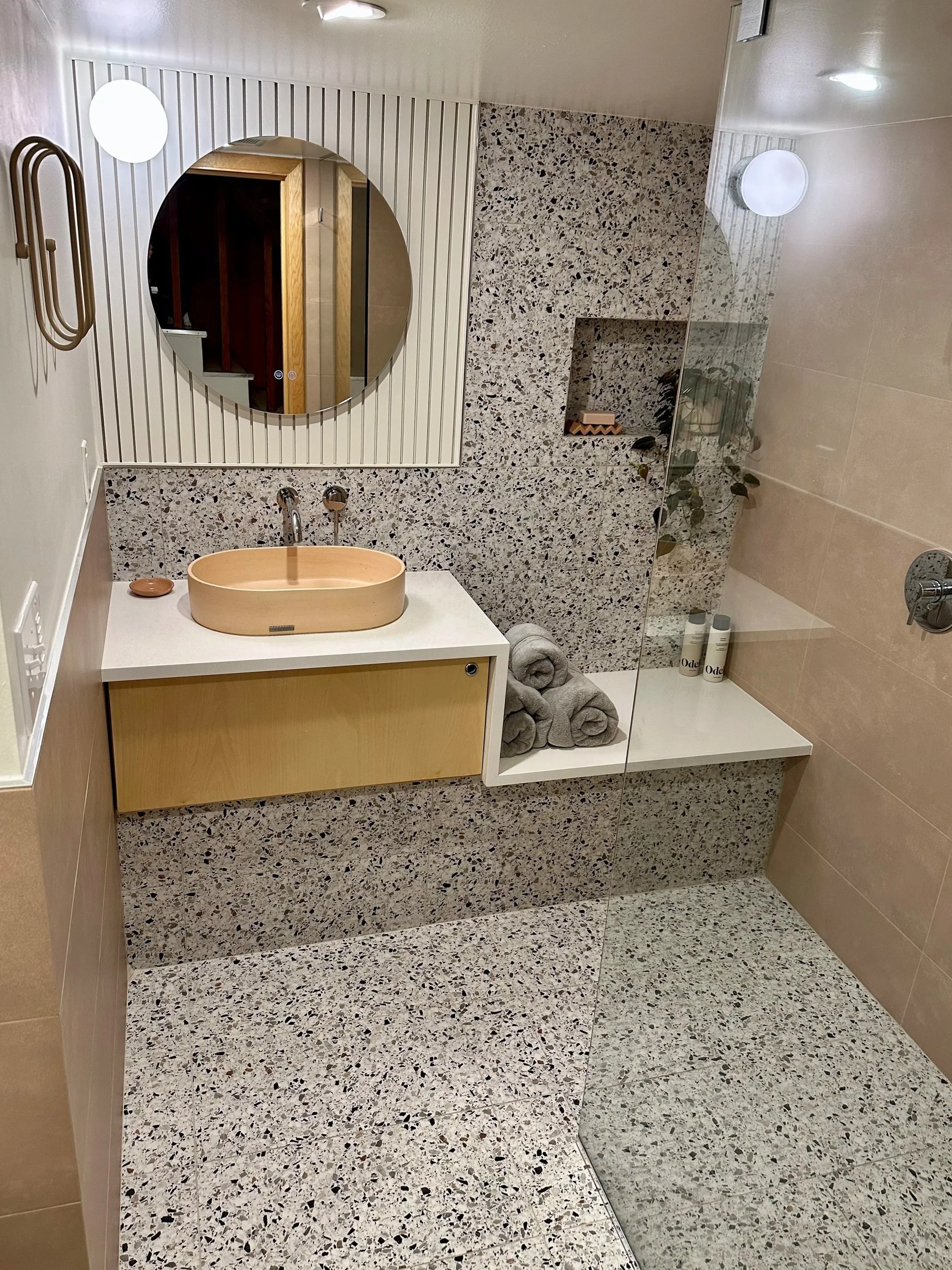 Modern bathroom with terrazzo walls and floor, a round mirror, a pink concrete sink bowl, rolled grey towels, and a glass shower area with beige tiles and a small shelf.