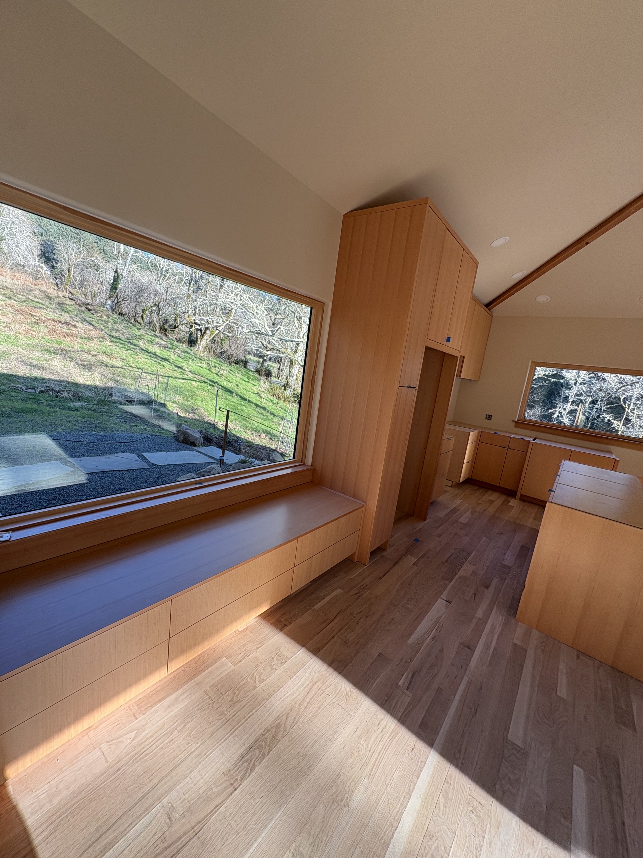 Empty room with large windows showing an outdoor view of trees and grass, wooden built-in furniture, hardwood floors, and ceiling with recessed lighting.