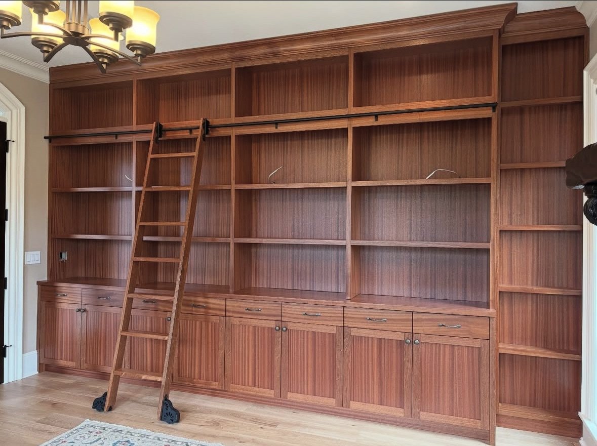 Empty built-in wooden bookshelf with a ladder in front of it, located in a room with hardwood floors and a chandelier ceiling light.