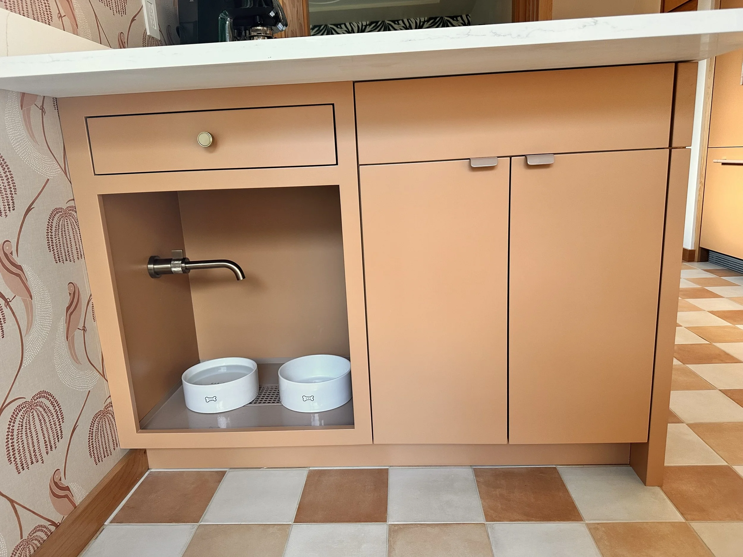 A kitchen counter with a built-in dog water dispenser featuring two bowls, a faucet, and a small vent, with peach-colored cabinets below and a white countertop above.