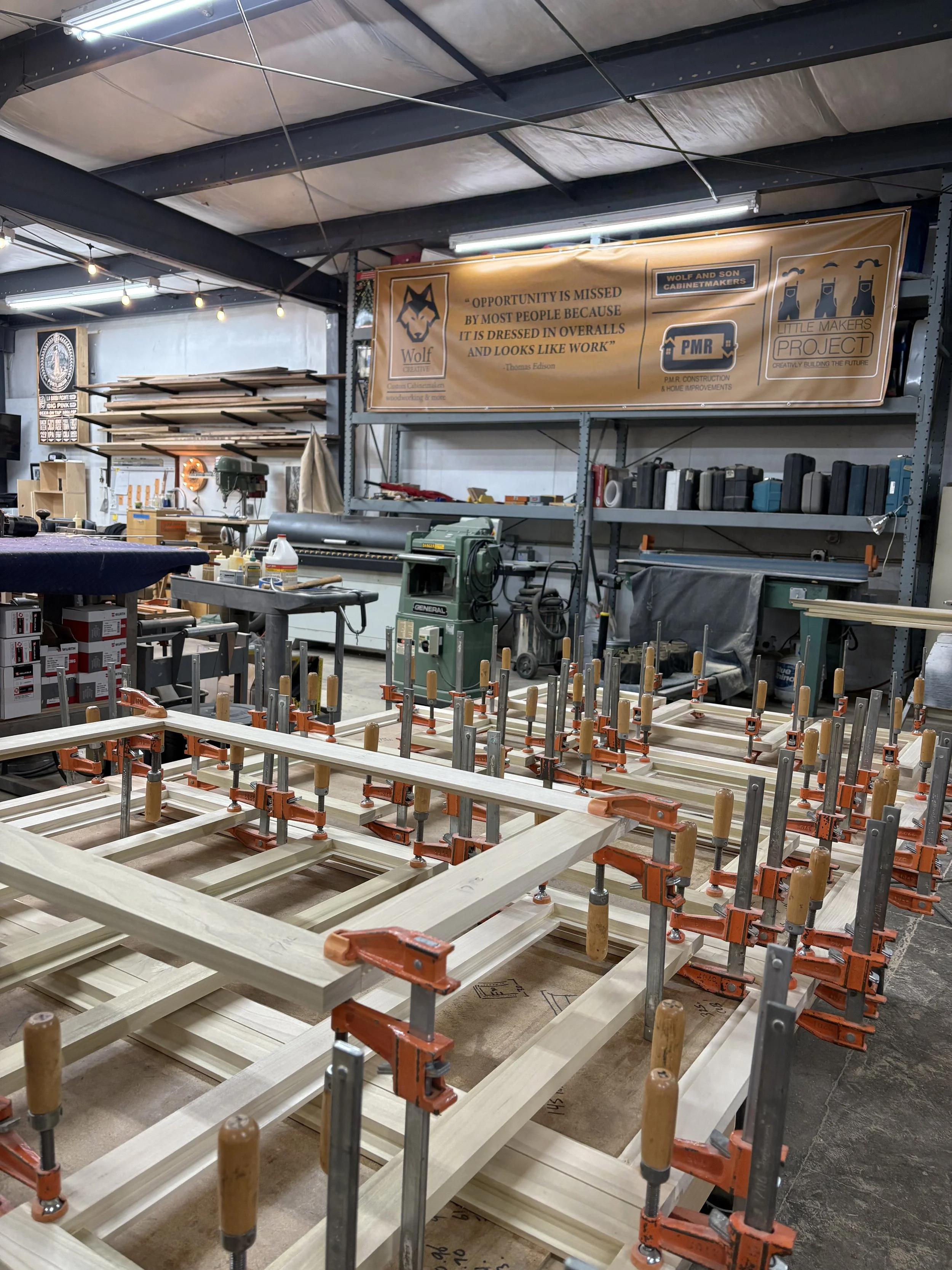 Woodworking shop with clamps securing wooden frames, shelves filled with tools and materials, and a banner with a wolf logo and quotes in the background.
