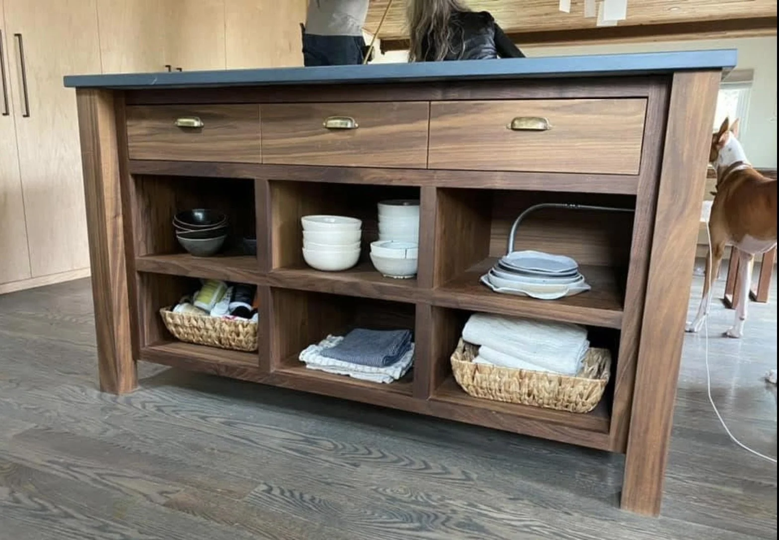 A wooden sideboard with three top drawers and open shelves below, containing bowls, plates, towels, and baskets, situated on a hardwood floor.