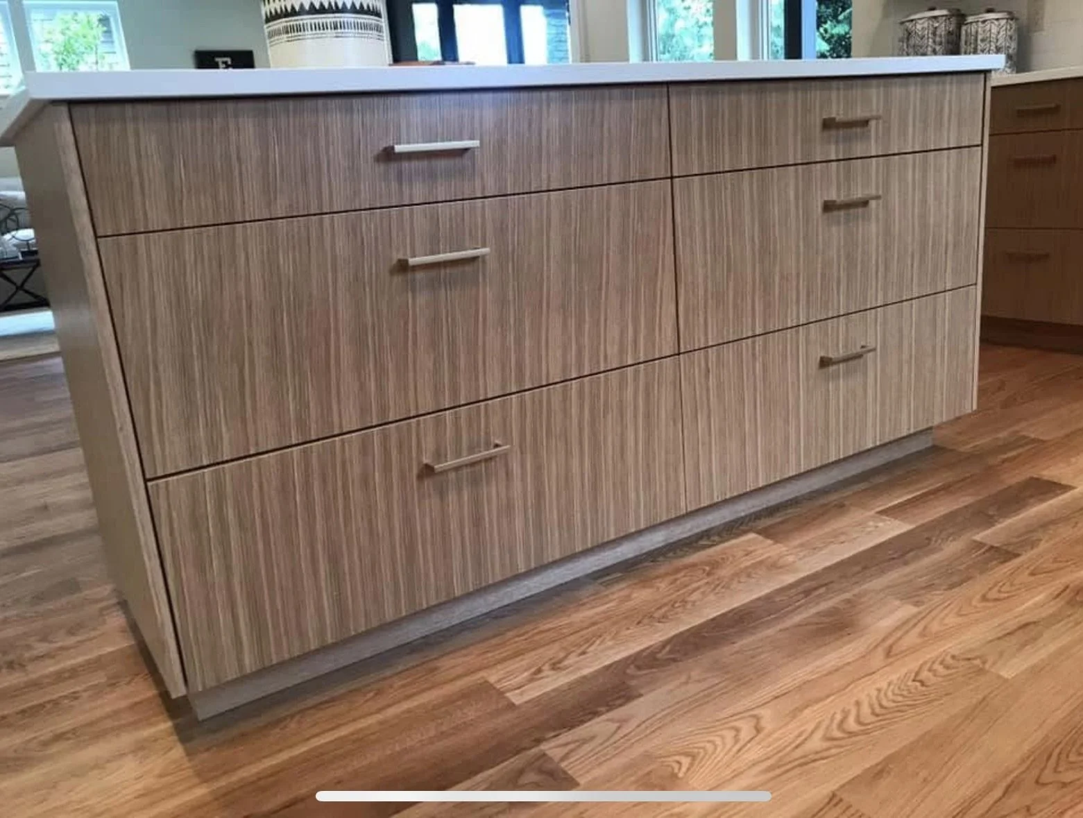 A wooden kitchen island with six drawers and silver handles, situated on a hardwood floor with a natural grain pattern in a well-lit room.