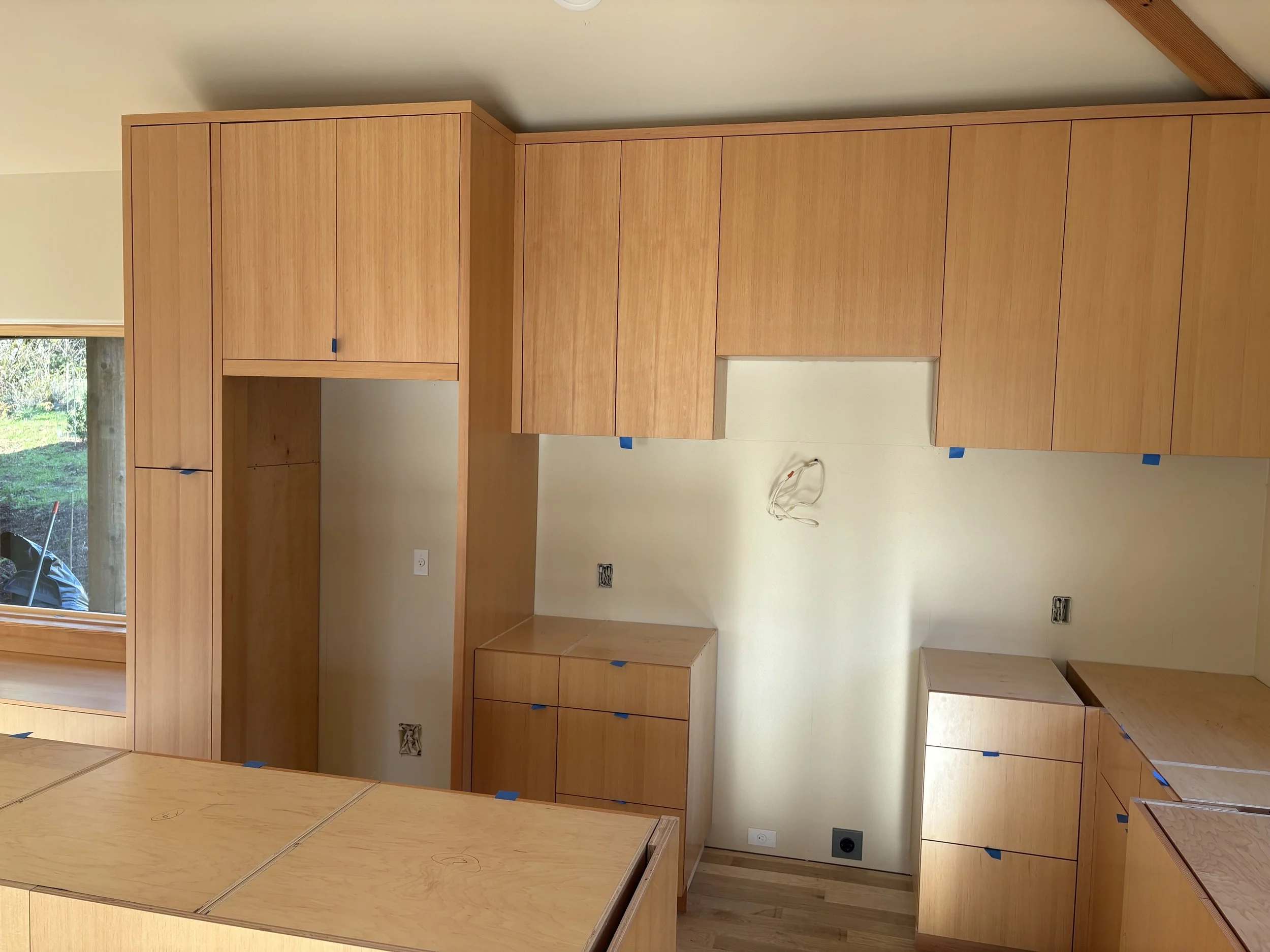 Kitchen cabinets being installed in a new home, with some blue painter's tape visible on the cabinets and electrical outlets without covers.