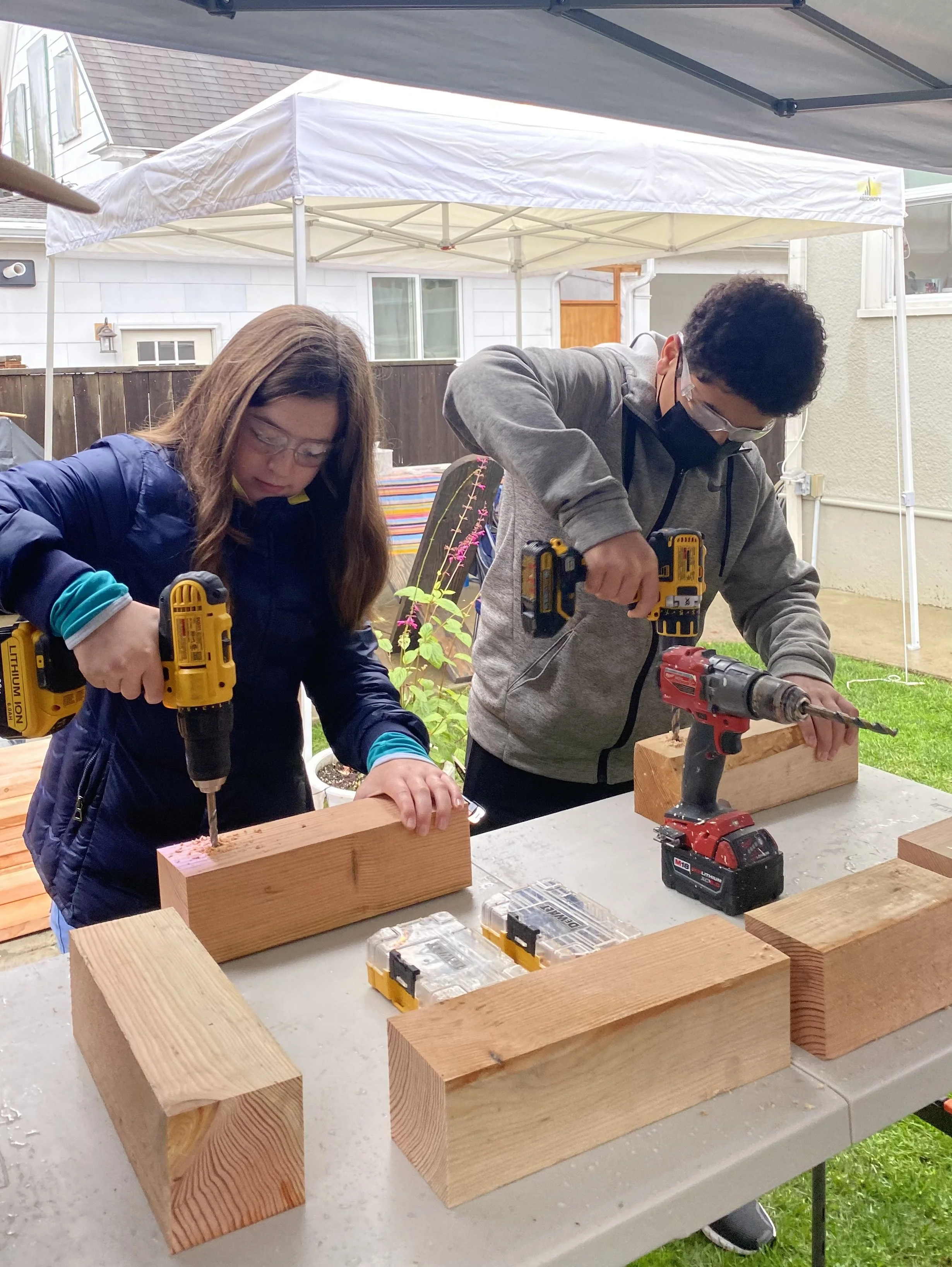 Two people working together on a woodworking project outdoors under a canopy, using power drills to screw together wooden beams.