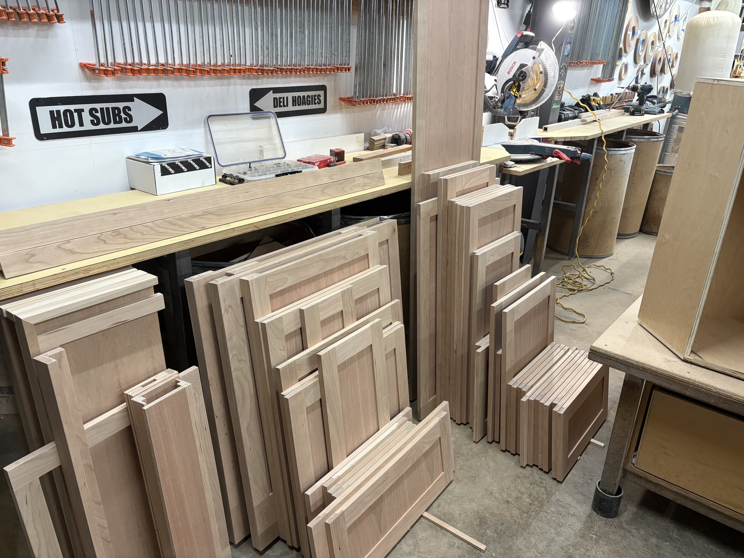 Lumber and woodworking tools in a workshop, with stacked wooden cabinet doors, a chop saw, and storage containers.