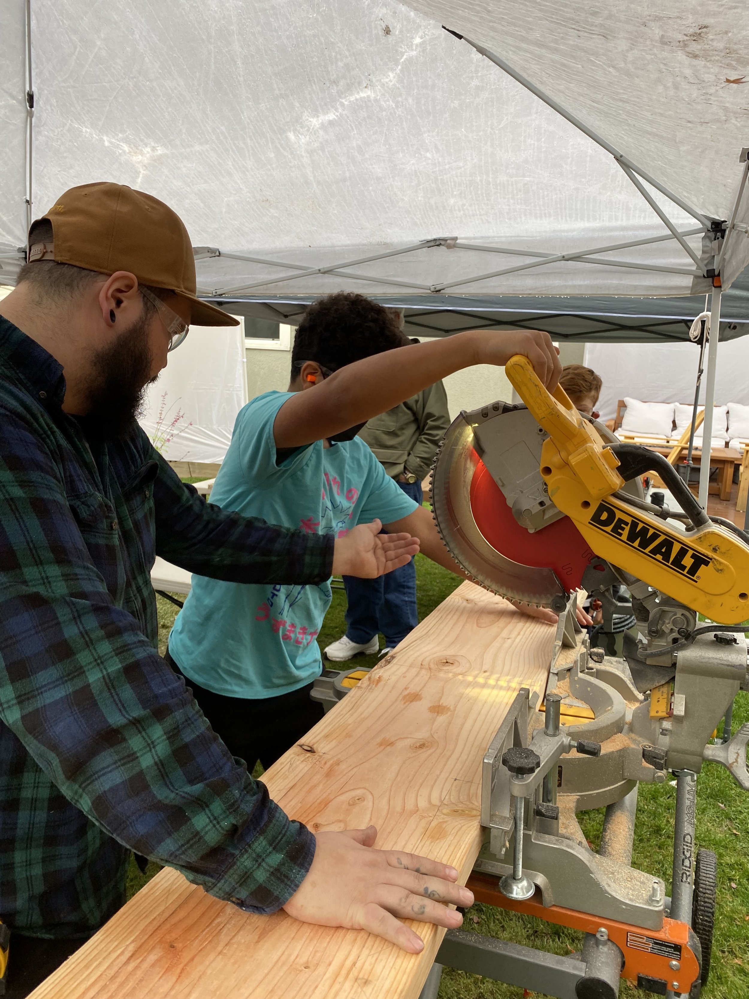 A man and a child working together on a wood-cutting project using a yellow Dewalt miter saw under a tent, with people and outdoor furniture in the background.