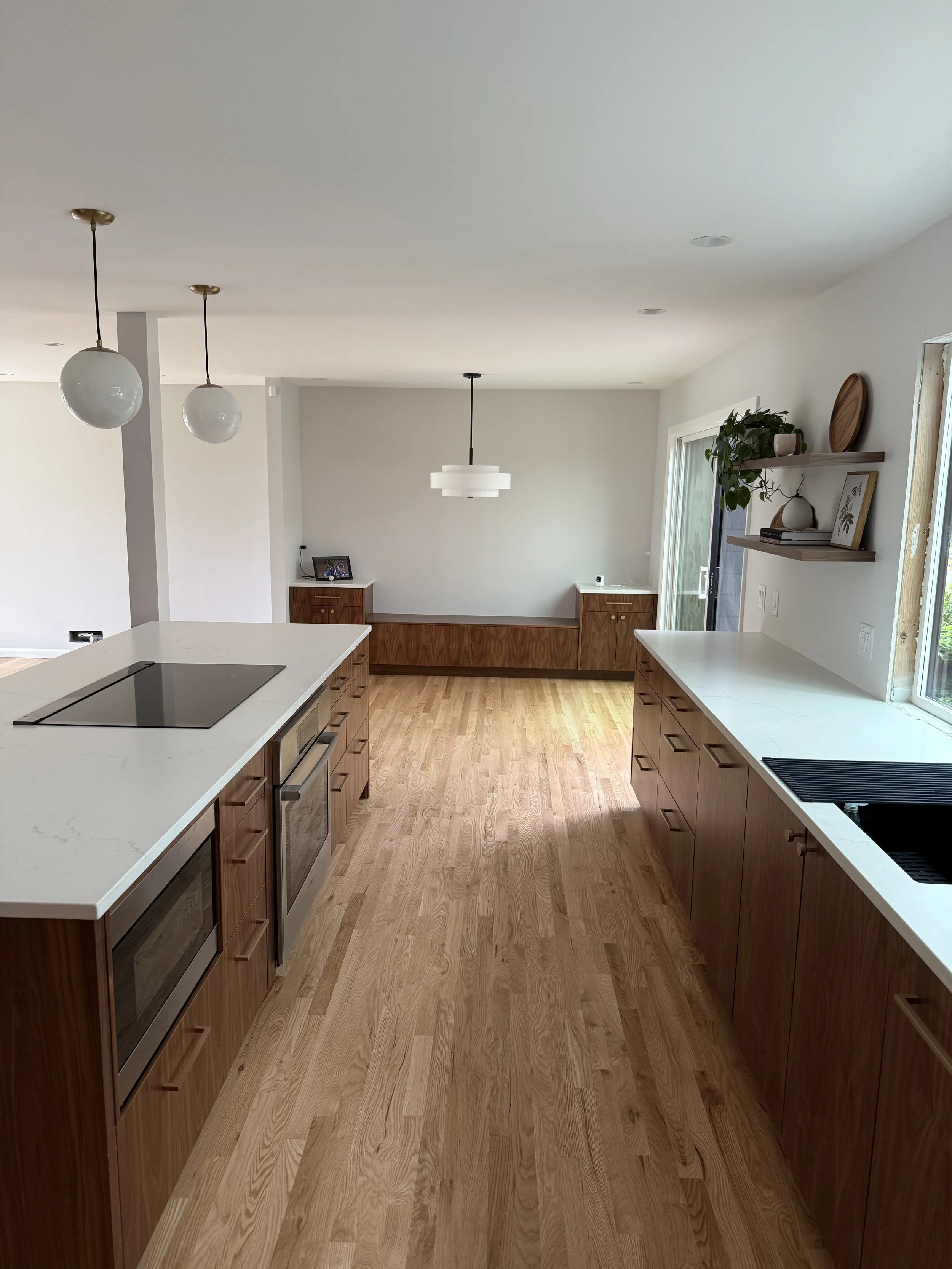 Modern kitchen with white countertops, wooden cabinets, and hardwood floors. Pendant and ceiling lights, open window, and floating shelves with decor.
