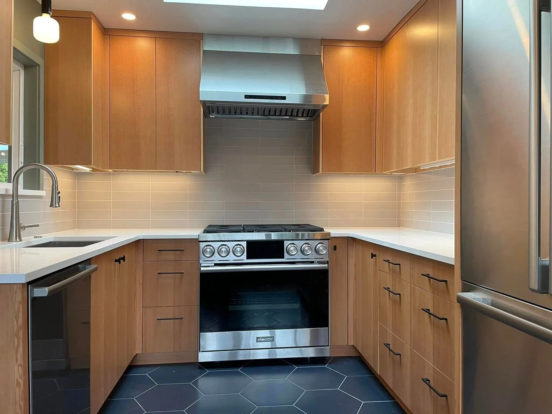 Modern kitchen with wooden cabinets, a stainless steel stove and range hood, black hexagonal floor tiles, white countertops, and a tan tile backsplash.