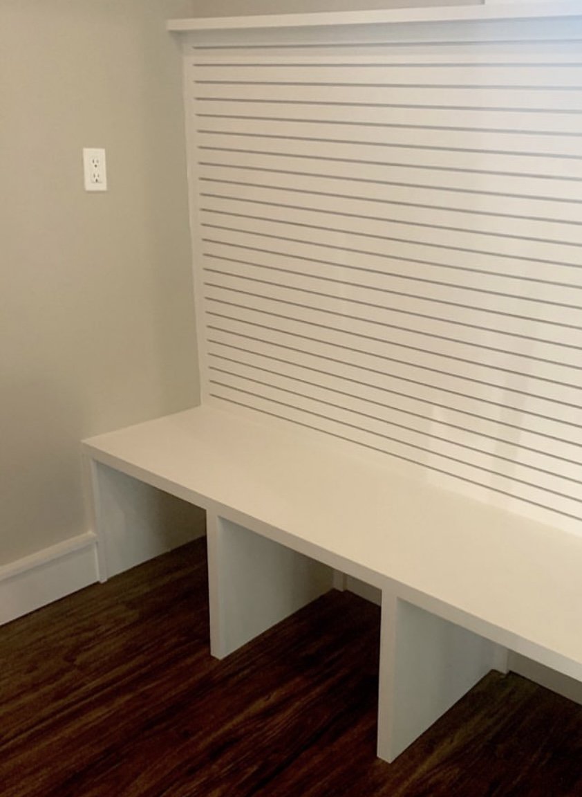Empty white built-in desk with three compartments underneath, against a wall with horizontal white slat paneling, light-colored walls, and dark wood flooring.