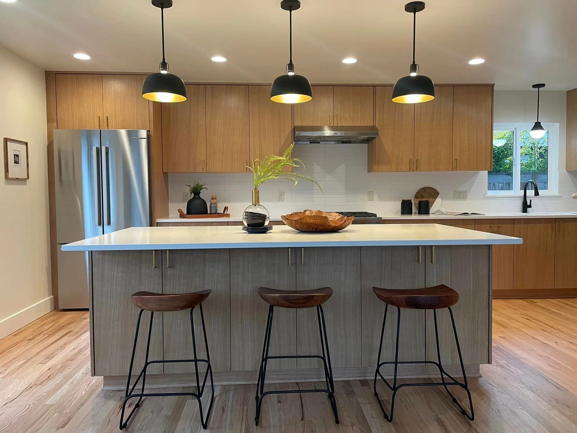 Modern kitchen with a large island, wooden cabinets, stainless steel refrigerator, black pendant lights, and wooden bar stools.
