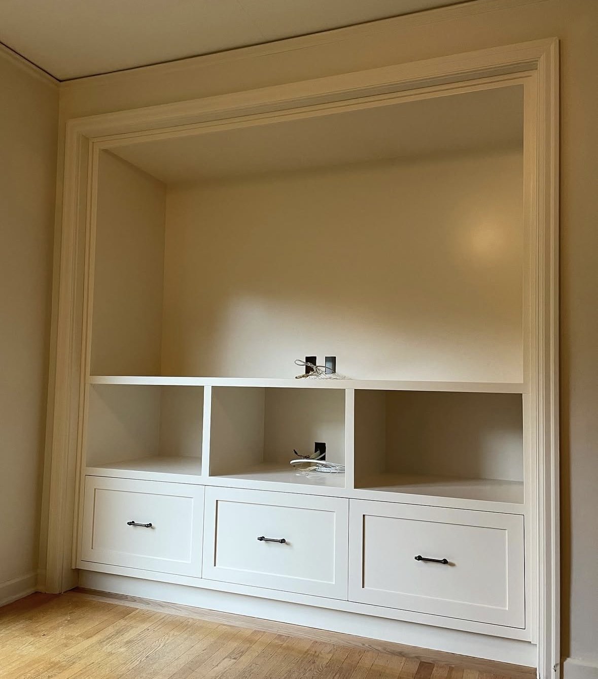 Empty built-in white wooden cabinet with three drawers at the bottom and open shelves above, located in a room with wooden floor and beige walls.
