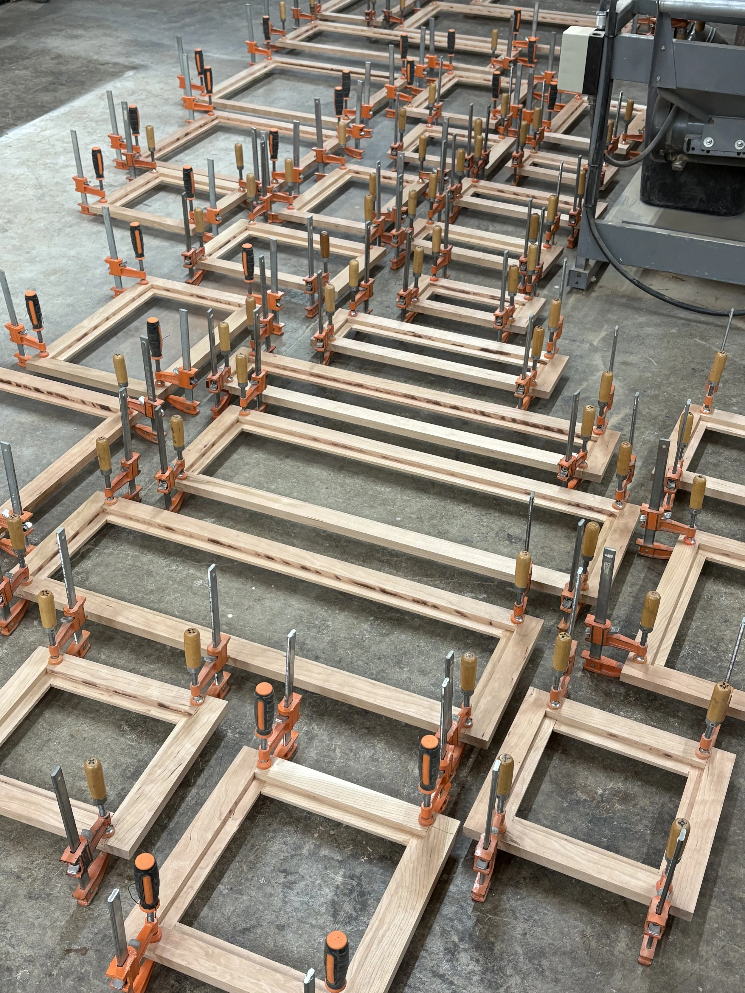 Numerous wooden frames clamped together with orange and silver clamps, arranged on the floor in a woodworking workshop.
