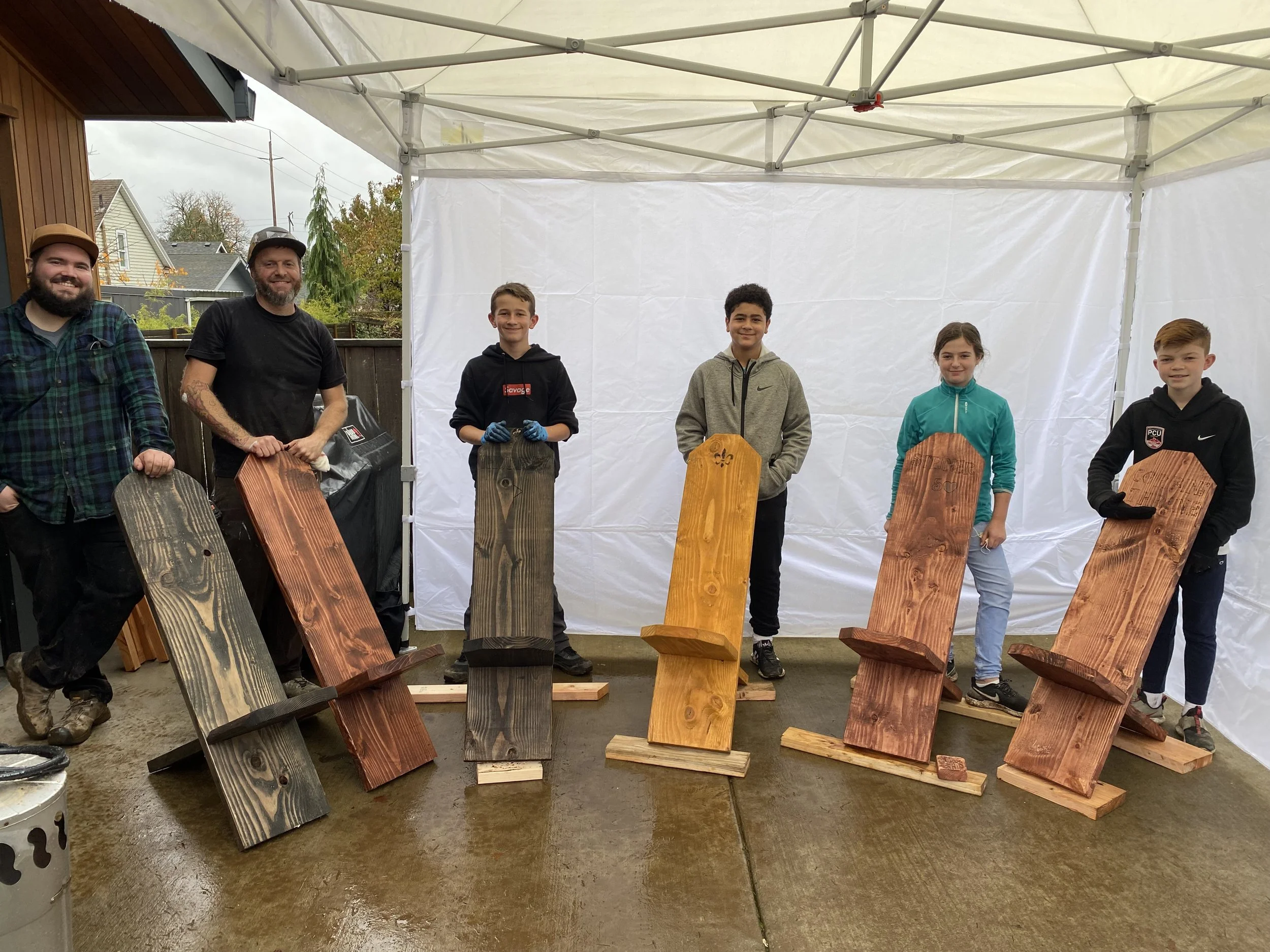Group of five people, two adults and three children, standing under a canopy, holding finished wooden sleds on a wet outdoor surface, with houses and trees in the background.