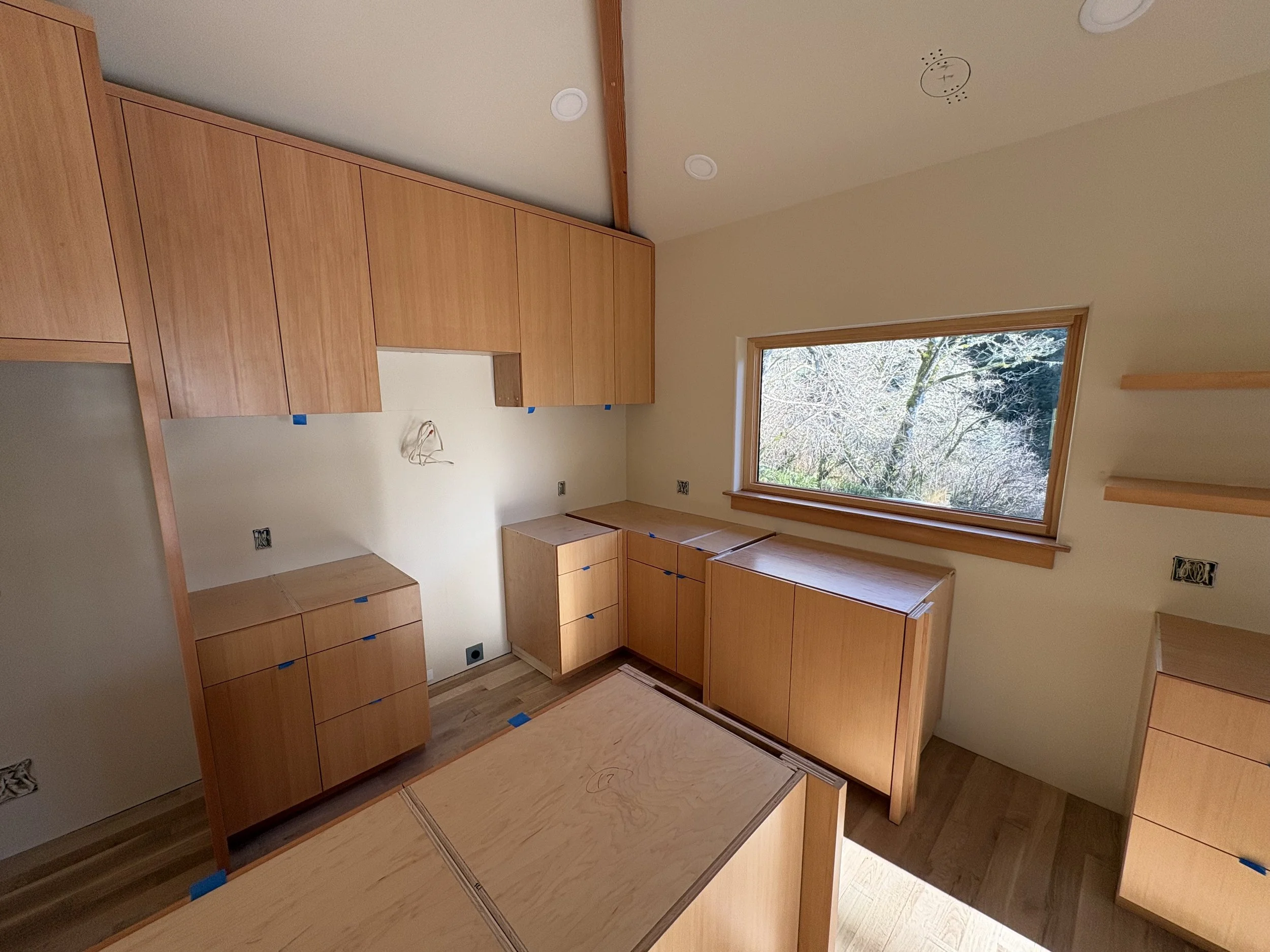 Empty kitchen with natural wood cabinets and drawers, a large window overlooking trees, and partially installed electrical outlets.
