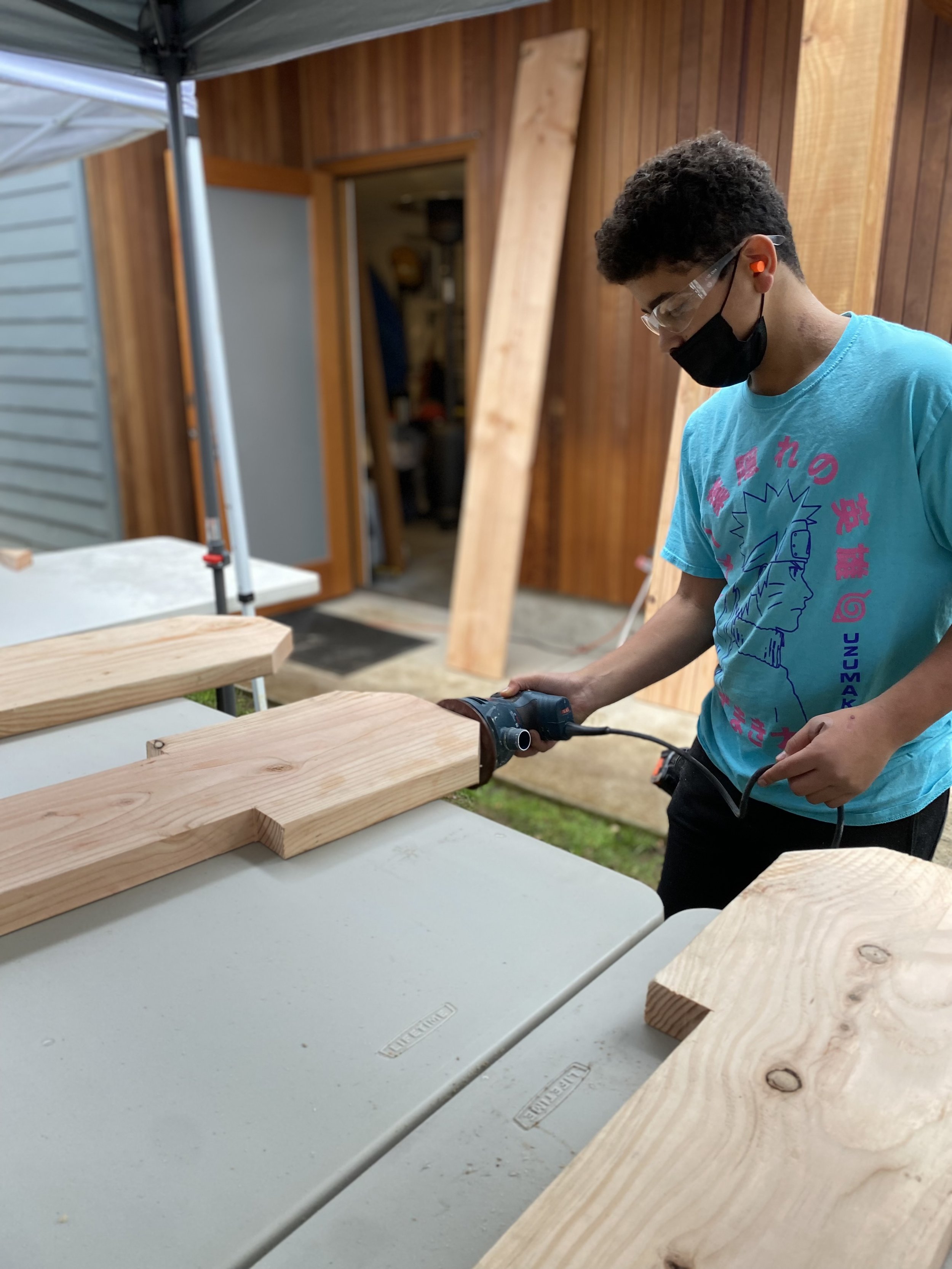 A person wearing a face mask, safety glasses, and ear protection uses an electric saw to cut a piece of wood on a workbench in a woodworking shop or outdoor workspace.
