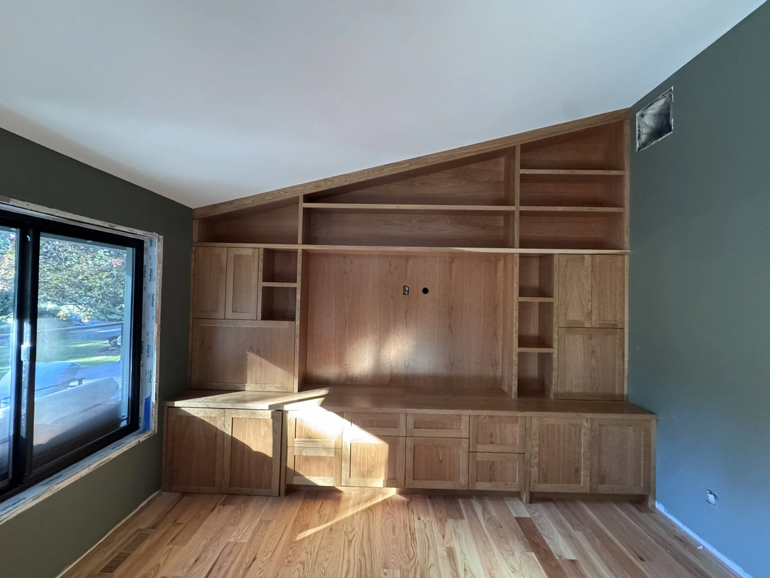 A room with a large built-in wooden bookshelf and cabinets against an angled wall, next to a window showing a view of trees and a street outside.