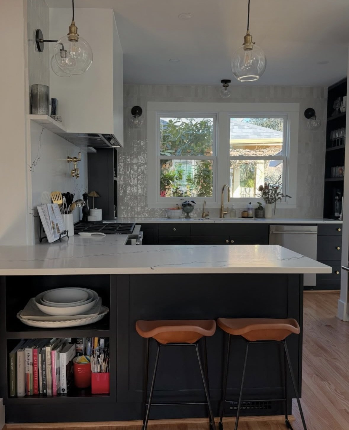 Modern kitchen with white marble countertops, dark cabinetry, black shelves, and two eating stools at the island. Large window over the sink lets in natural light, and there are glass pendant lights hanging from the ceiling.
