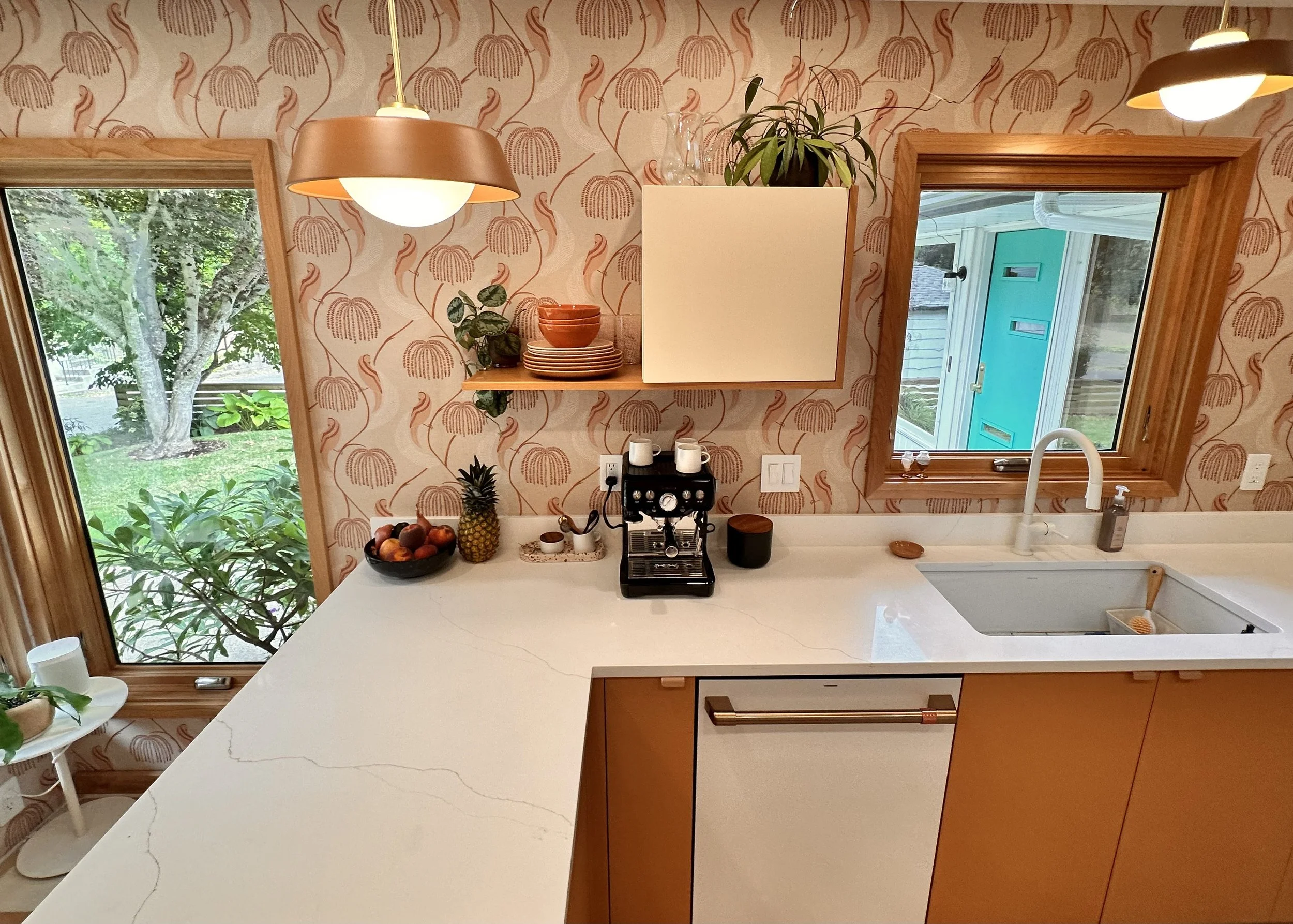 A modern kitchen with a marble countertop, fruit basket with pineapple, apples, bananas, and peaches, coffee machine, wall-mounted shelf with dishes and plants, two windows showing trees and a neighbor's house, and wooden accents.