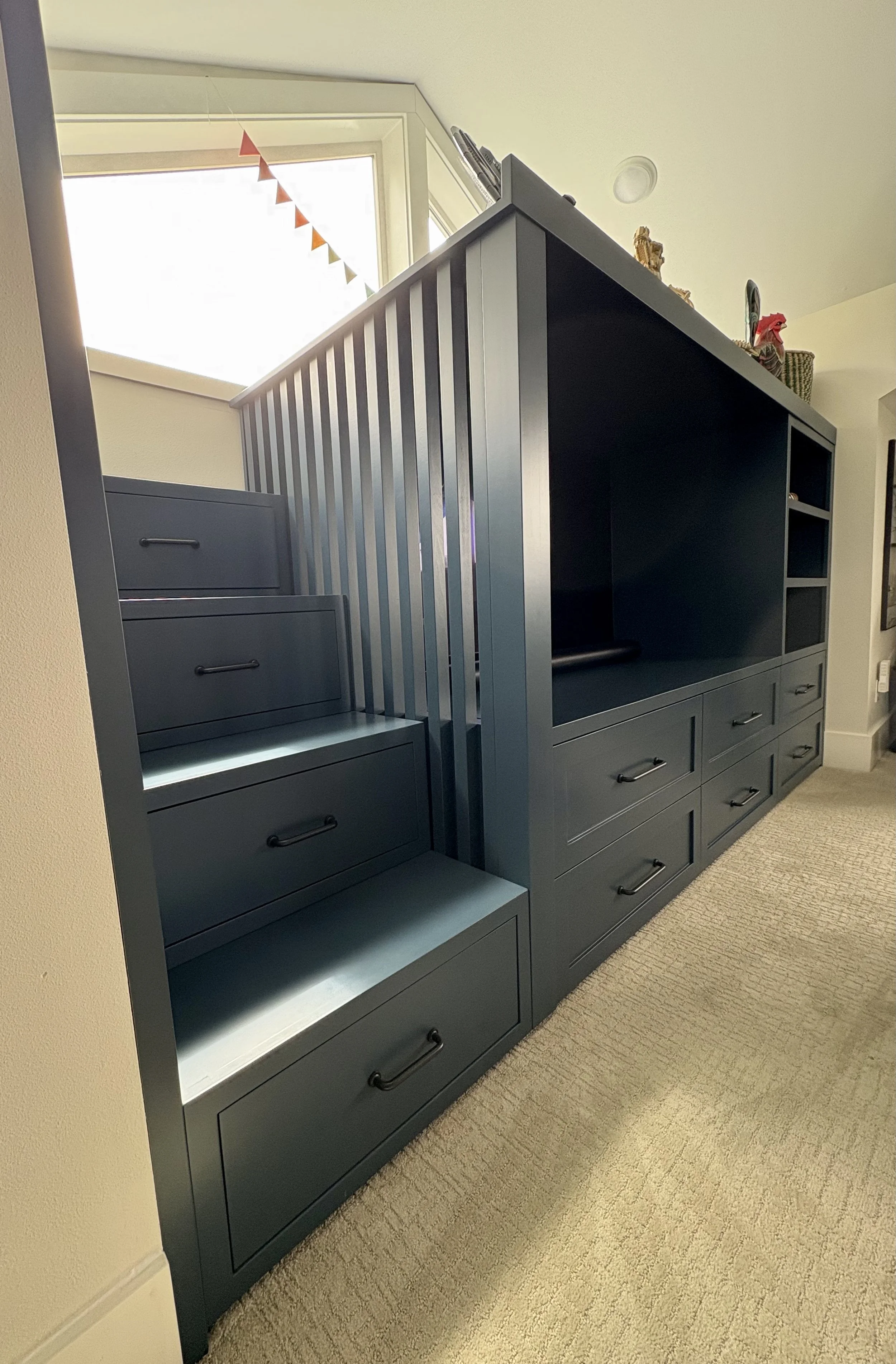 Blue built-in cabinetry with drawers and open shelves next to a staircase with a metal balustrade, near a window with triangular bunting hanging from the top.