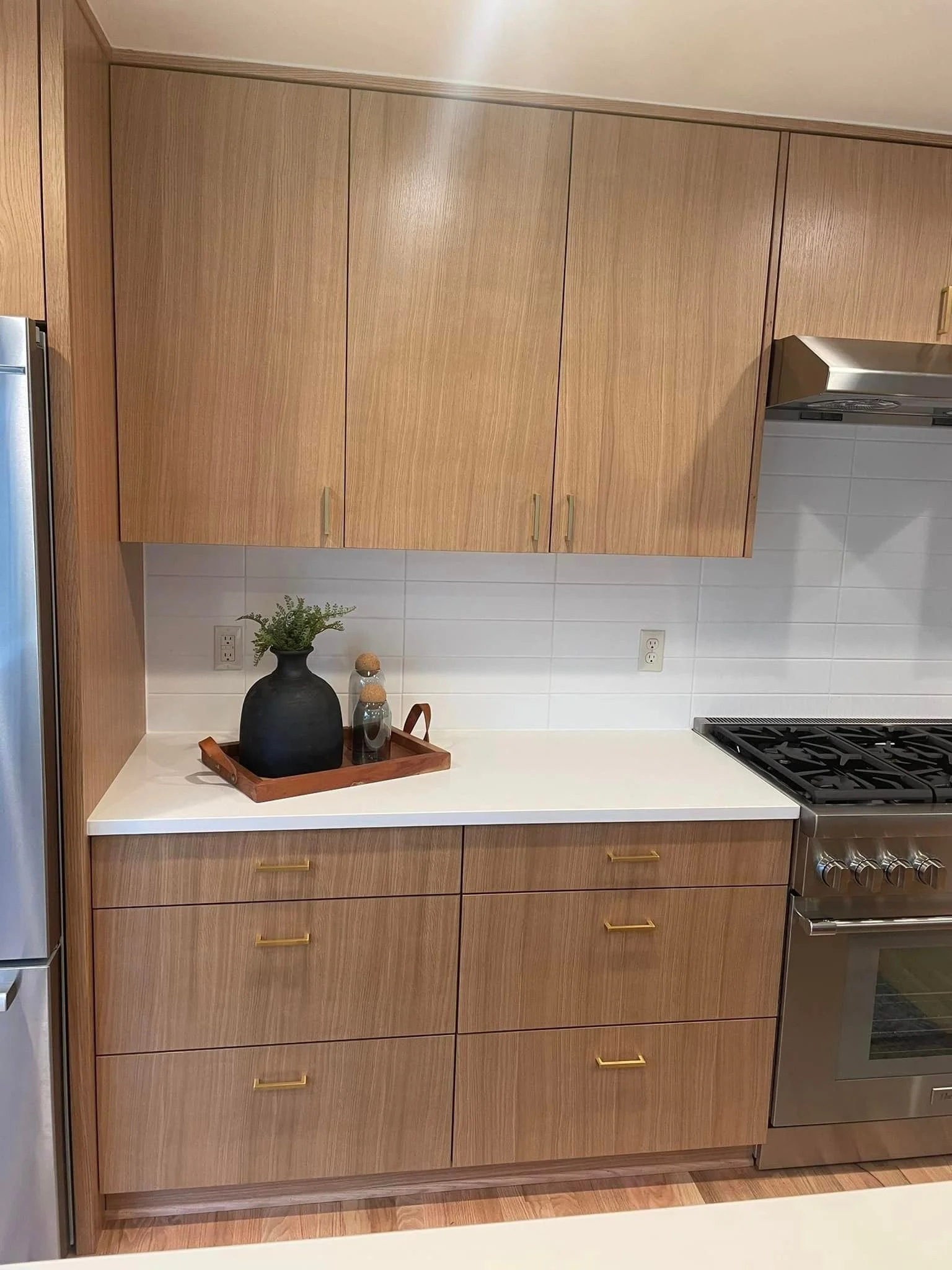 Kitchen with wooden cabinets, white countertop, black vase with green plant, tray with small objects, and stainless steel stove.