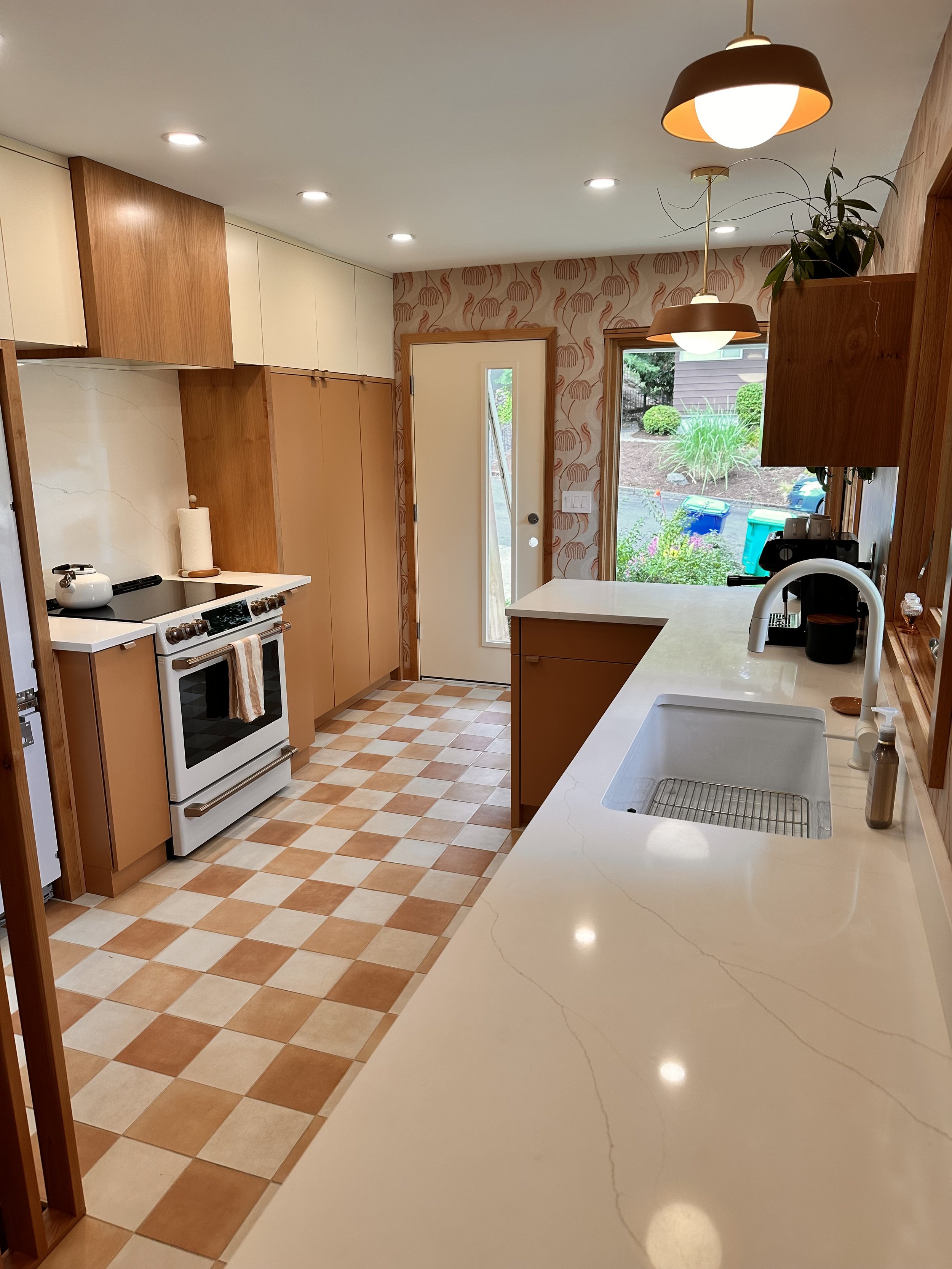 Kitchen with checkered tan and white floor tiles, beige countertops, wooden cabinets, a white oven, and a window overlooking a garden with plants. Modern ceiling lights and a door leading outside.