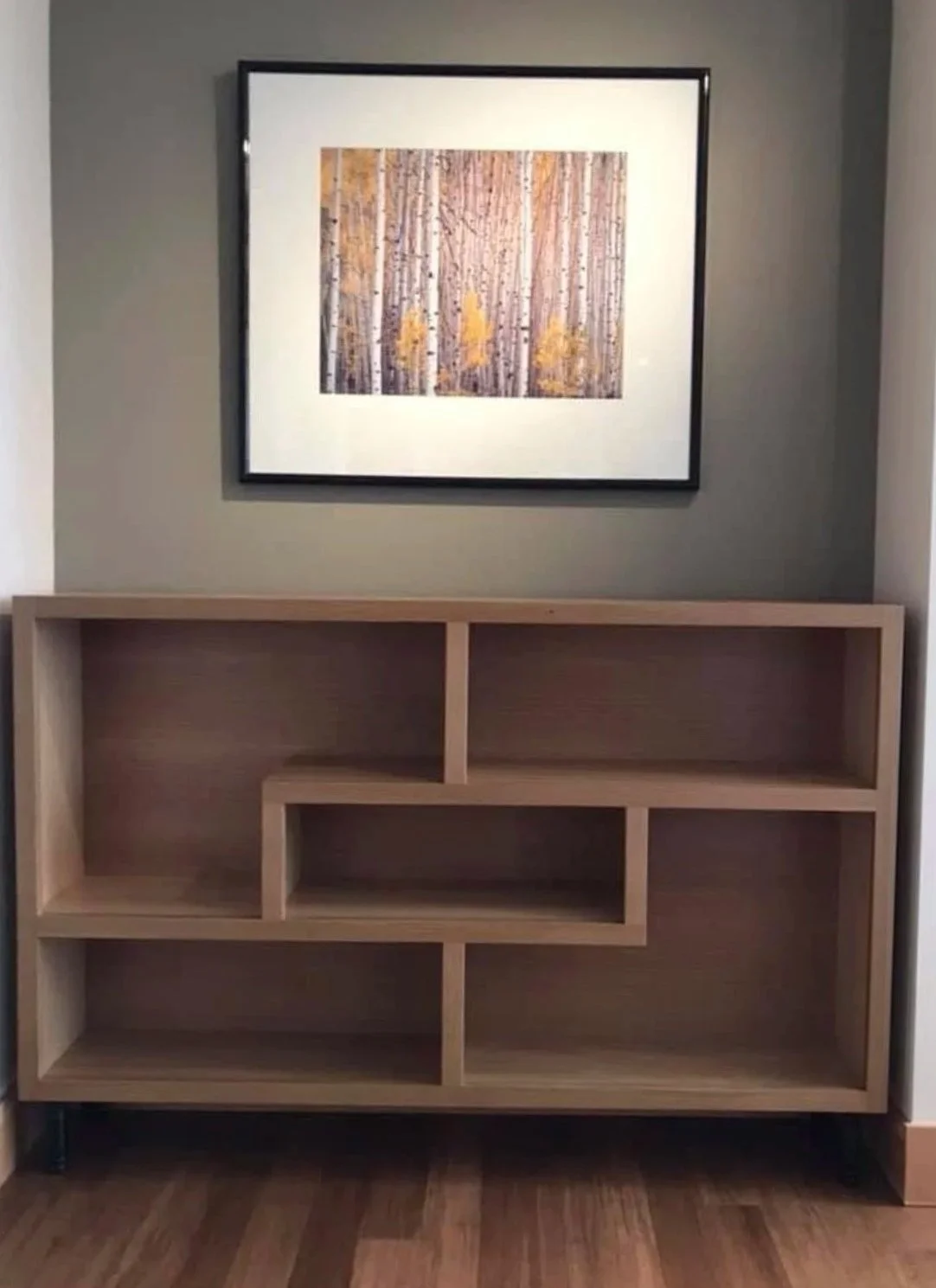 Empty wooden bookshelf with geometric compartments in front of a gray wall, above a framed landscape painting of a forest with yellow leaves.